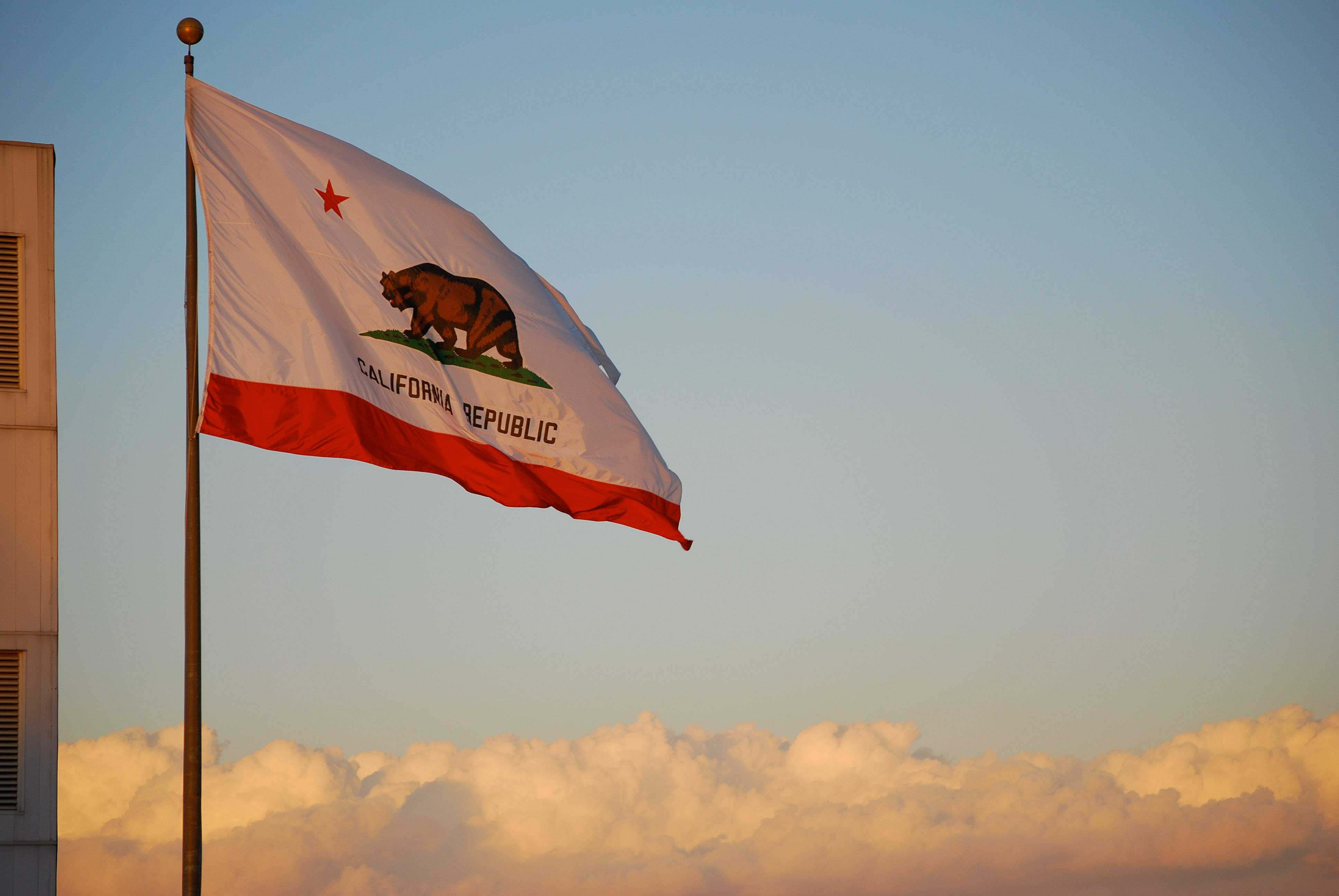 Flag 桌布 壁紙 — 加州州旗在柔和的金色日落天空下自豪地飄揚，下方有蓬鬆的雲朵。