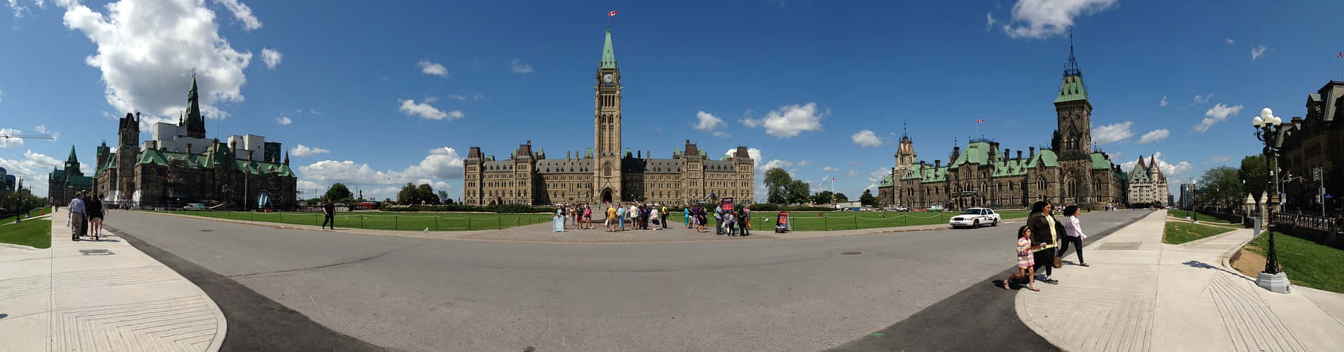 Panoramic 桌布 壁紙 — 加拿大國會大廈的全景，在明亮的藍天和白雲下，遊客在寬闊的小徑上行走。