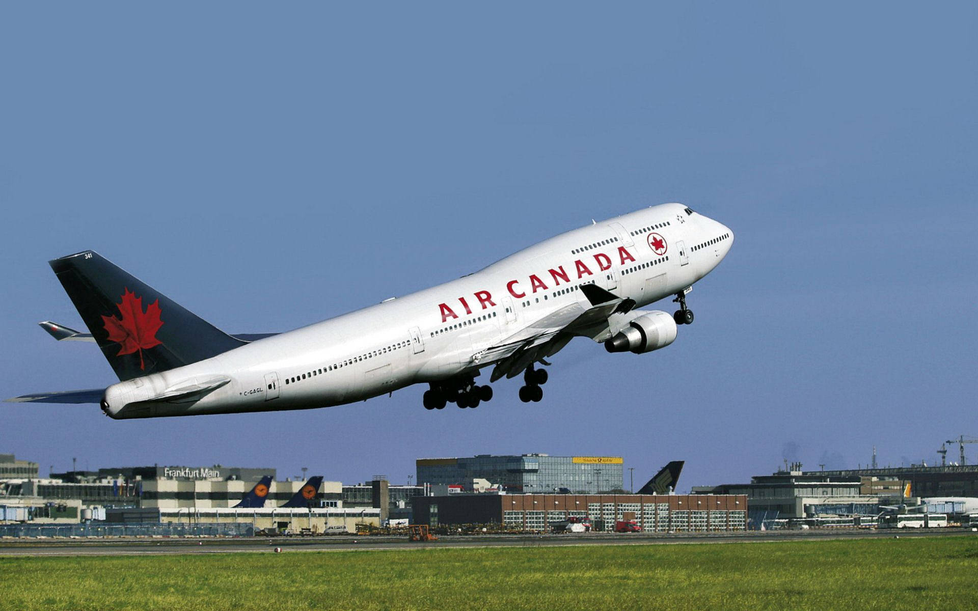 飛機 桌布 壁紙 — 加拿大航空波音747在晴朗藍天下起飛，下方有機場建築物和綠色草地。