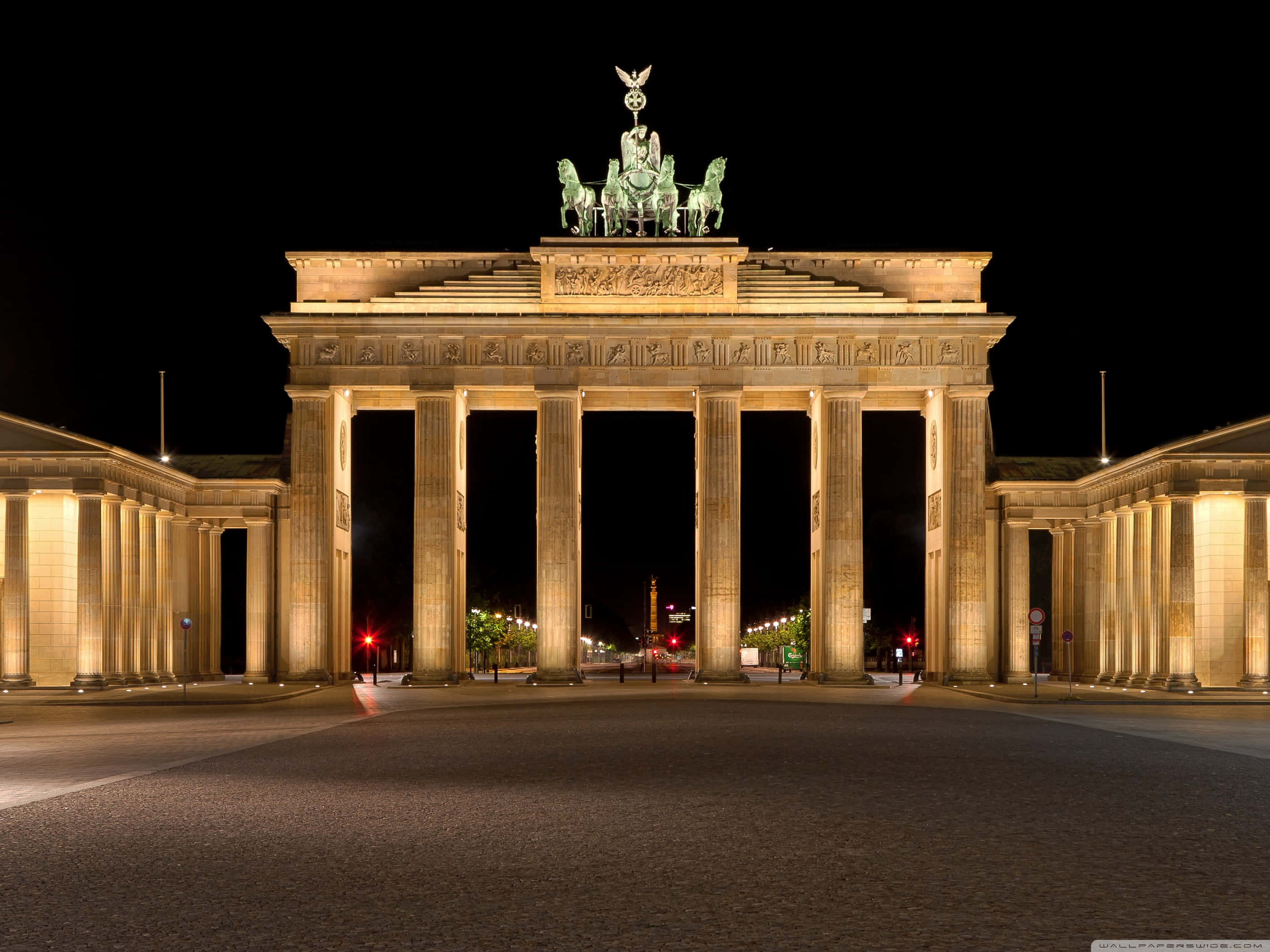 Berlin 桌布 壁紙 — 勃蘭登堡門在夜間被溫暖的燈光照亮，凸顯其新古典主義柱子和四馬戰車雕像，背景是黑暗的天空。