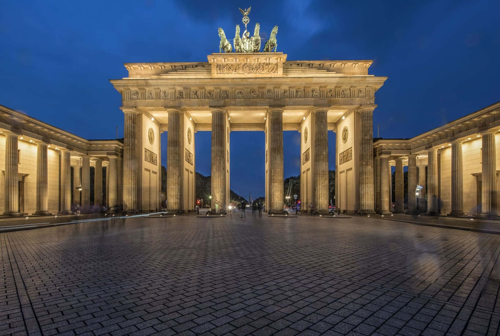 Berlin 桌布 壁紙 — 勃蘭登堡門在黃昏時亮起，溫暖的燈光映照在濕漉漉的鵝卵石上，深藍色的天空下。