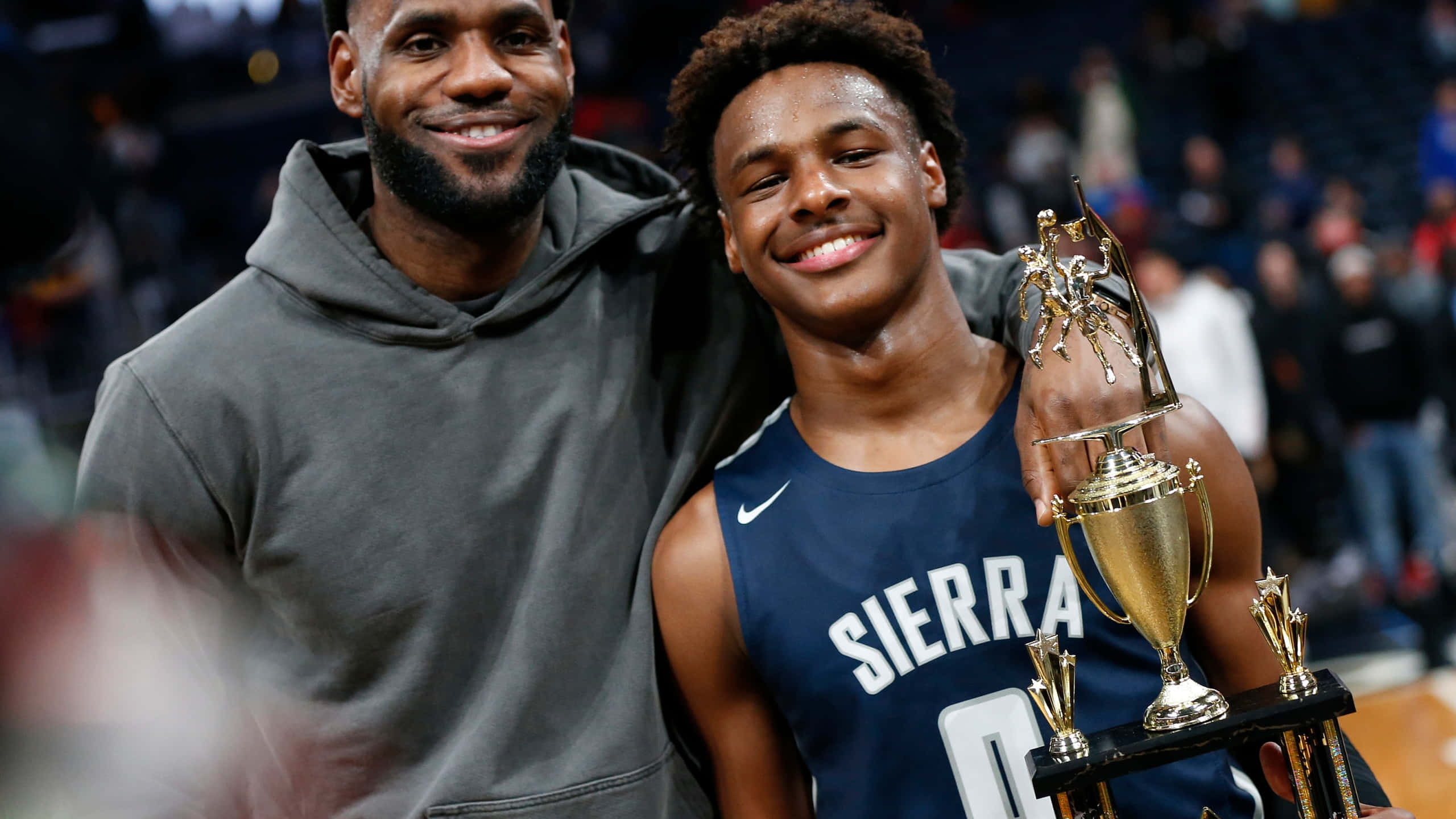 Bronny James 桌布 壁紙 — 勒布朗·詹姆斯在球場上與一位手持獎盃的年輕籃球運動員溫暖地微笑。