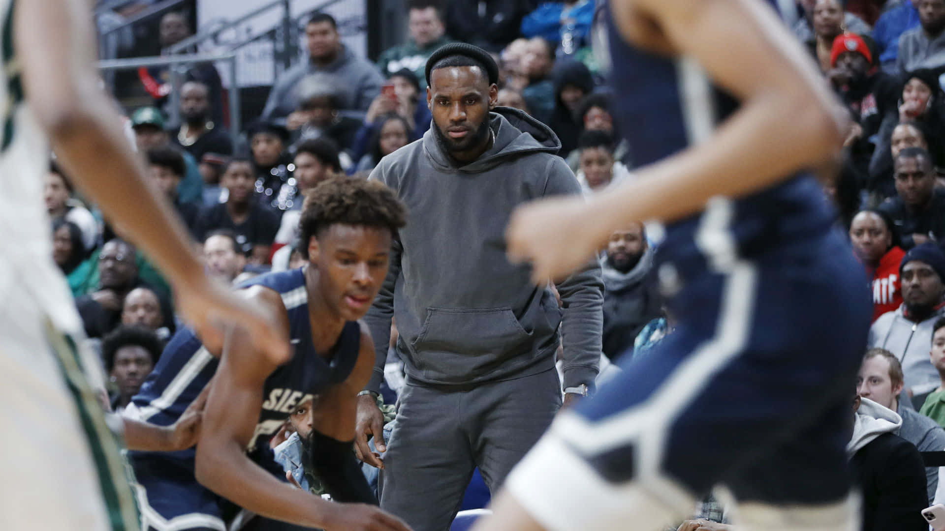 Bronny James 桌布 壁紙 — 勒布朗·詹姆斯在邊線專注地觀看，年輕籃球運動員在室內比賽中競技，觀眾歡呼喝采。