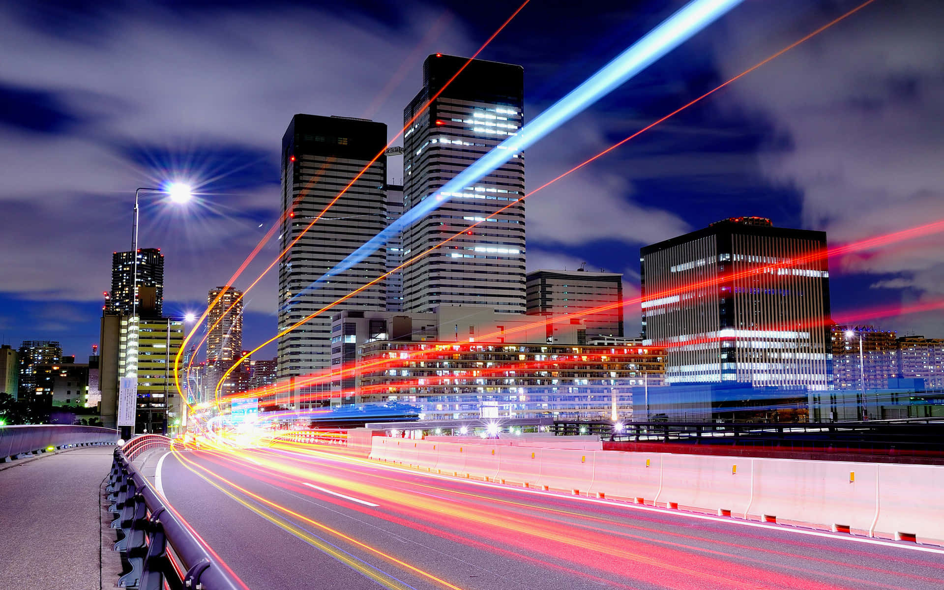夜空 桌布 壁紙 — 動感夜間城市景觀，車流光跡劃過高聳亮燈的摩天大樓，星空璀璨。