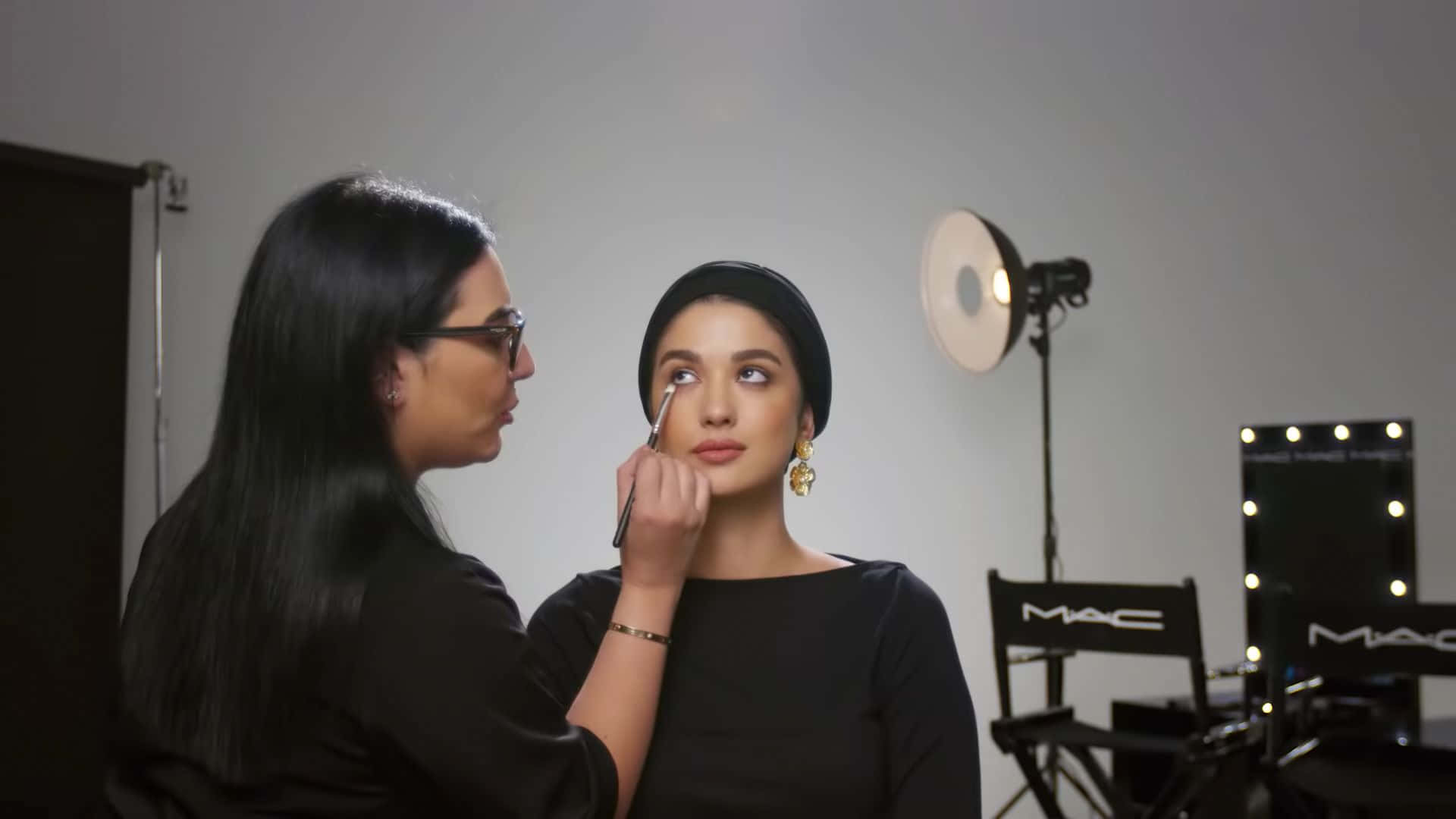 Mac Cosmetics 桌布 壁紙 — 化妝師在專業工作室中，在明亮的燈光和MAC品牌椅子下，為模特兒的眼睛畫眼線。