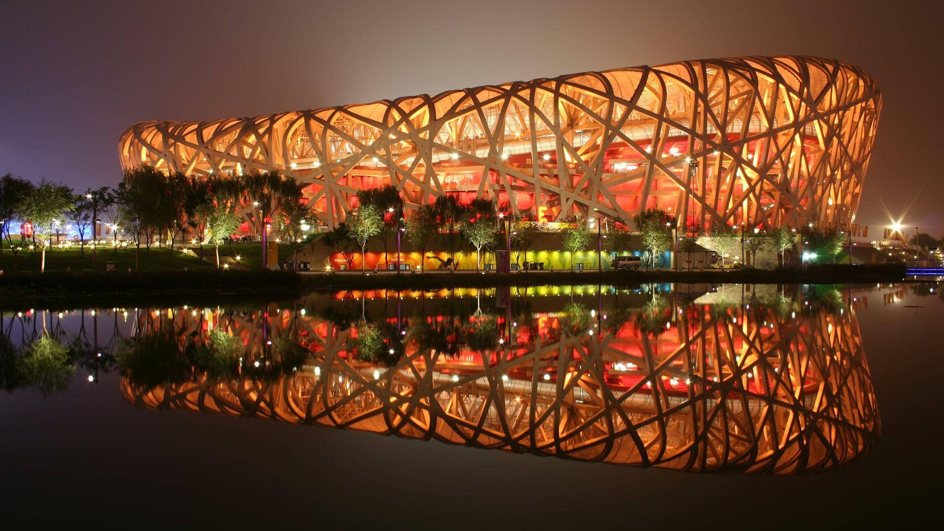Beijing 桌布 壁紙 — 北京國家體育場在夜晚溫暖地發光，其複雜的網格結構完美地倒映在下方平靜的水面上。