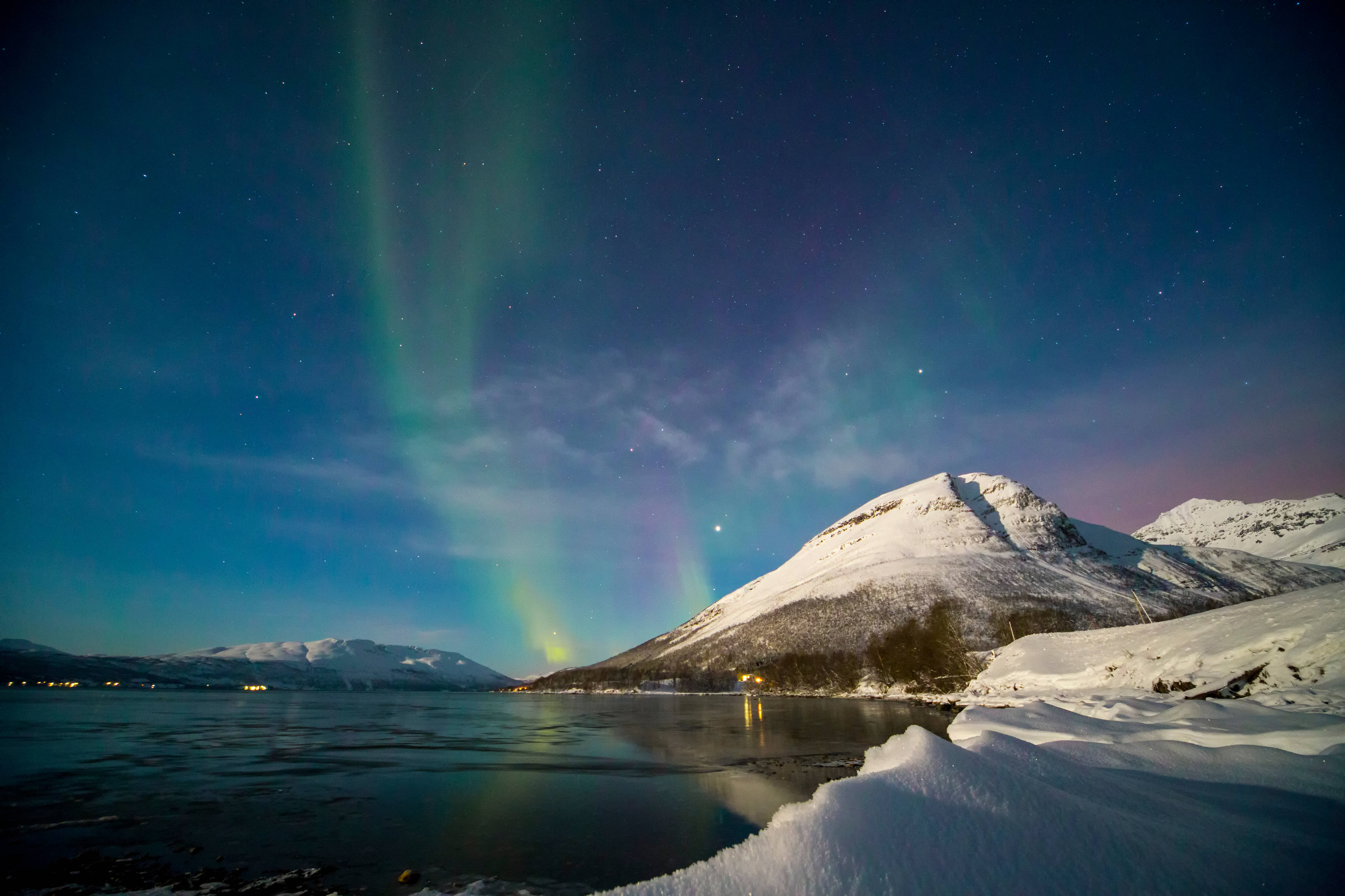 Norway 桌布 壁紙 — 北極光在綠色和紫色中舞動，映照在星空下冰凍湖面上的雪山之上。