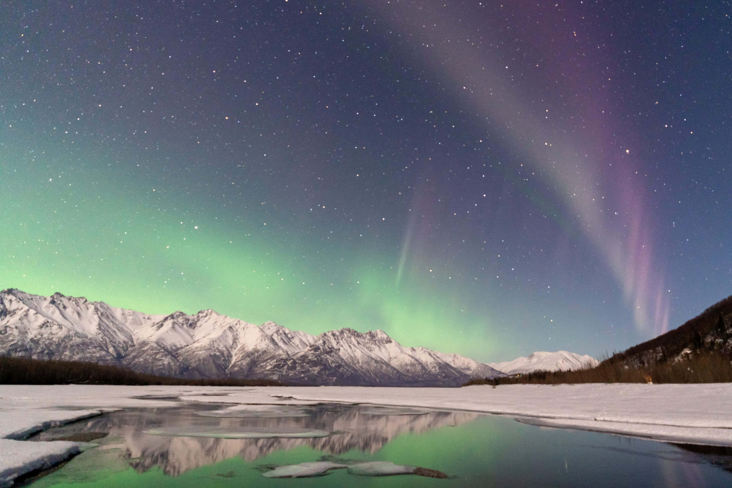 aurora borealis 桌布 壁紙 — 北極光在繁星點點的夜空中以鮮豔的綠色和紫色舞動，映照在結冰的湖面上的雪山之上。