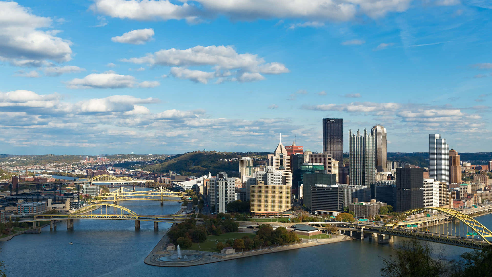 Pittsburgh Skyline 桌布 壁紙 — 匹茲堡天際線的全景，黃色橋樑橫跨河流，在明亮的藍天下有蓬鬆的雲朵。