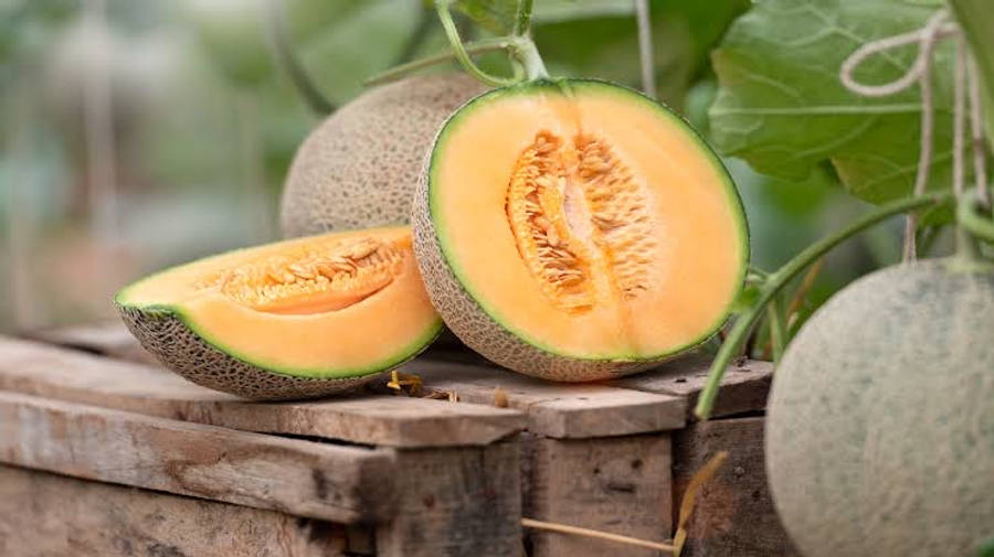 Fruit 桌布 壁紙 — 半個哈密瓜，橙色果肉和綠色外皮，放在一個風化的木箱上，旁邊有完整的哈密瓜和茂盛的綠葉。