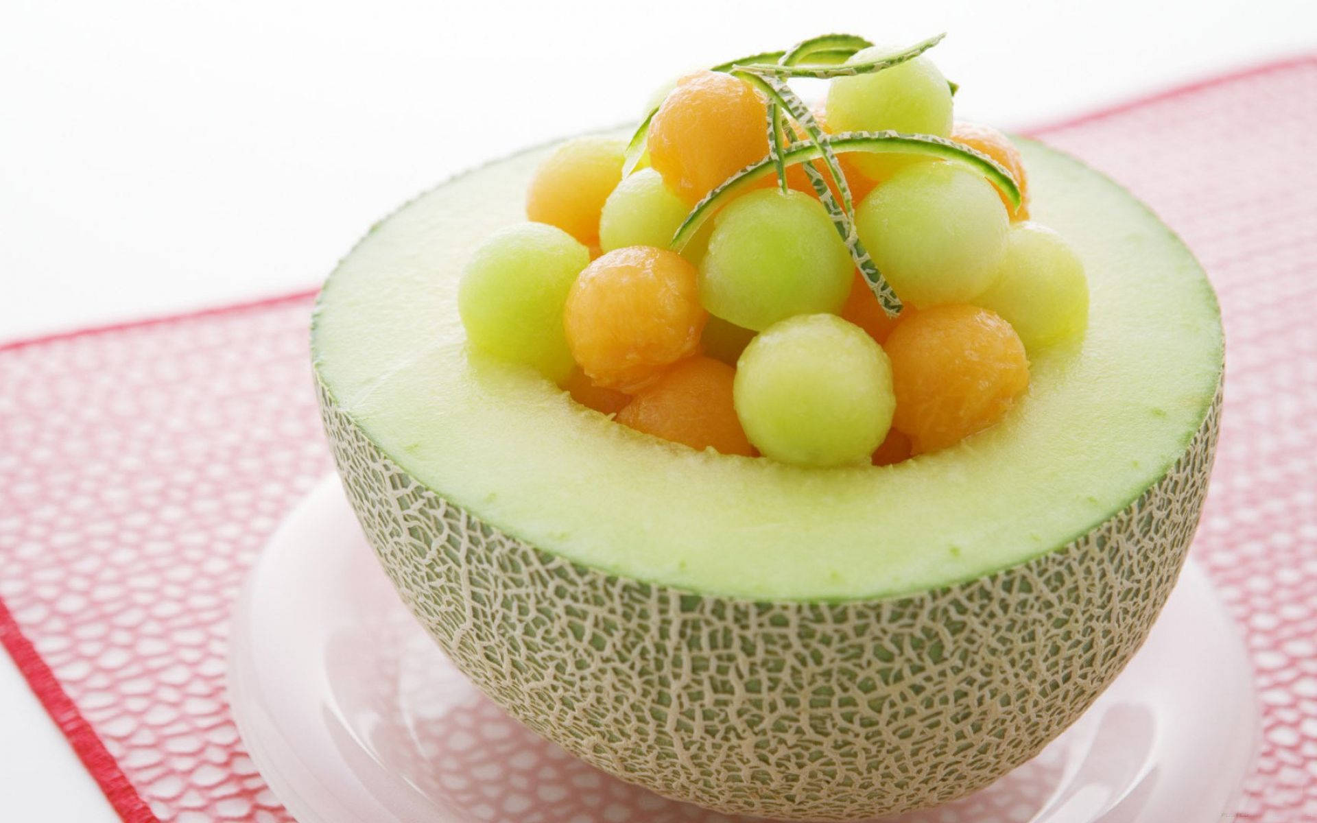 Fruit 桌布 壁紙 — 半個哈密瓜，裡面裝滿彩色蜜瓜球，並用迷迭香裝飾，放在粉紅色紋理墊上。