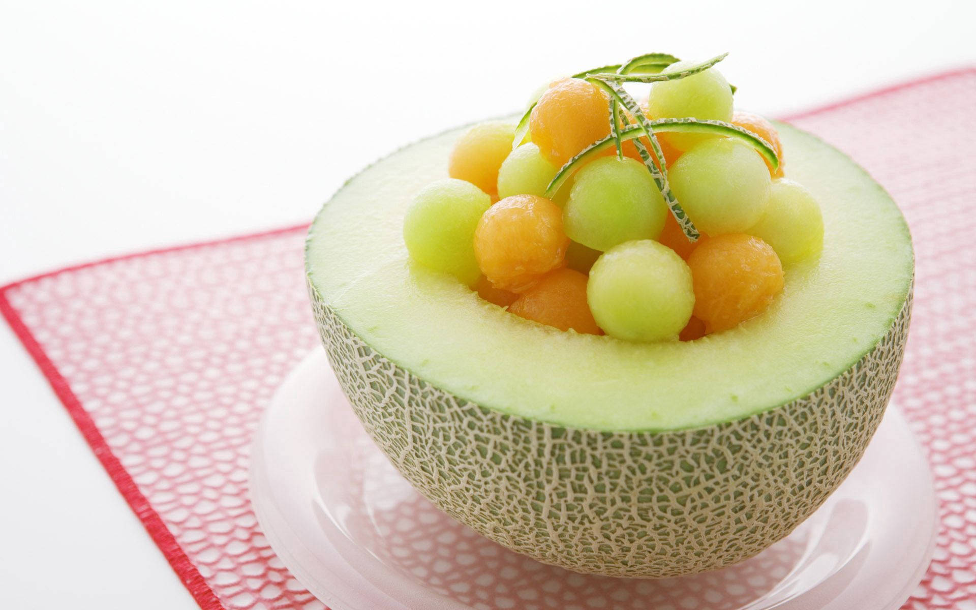 Fruits 桌布 壁紙 — 半個哈密瓜，裡面裝滿彩色蜜瓜球，並用一枝綠色香草點綴，放在有圖案的紅色墊子上。