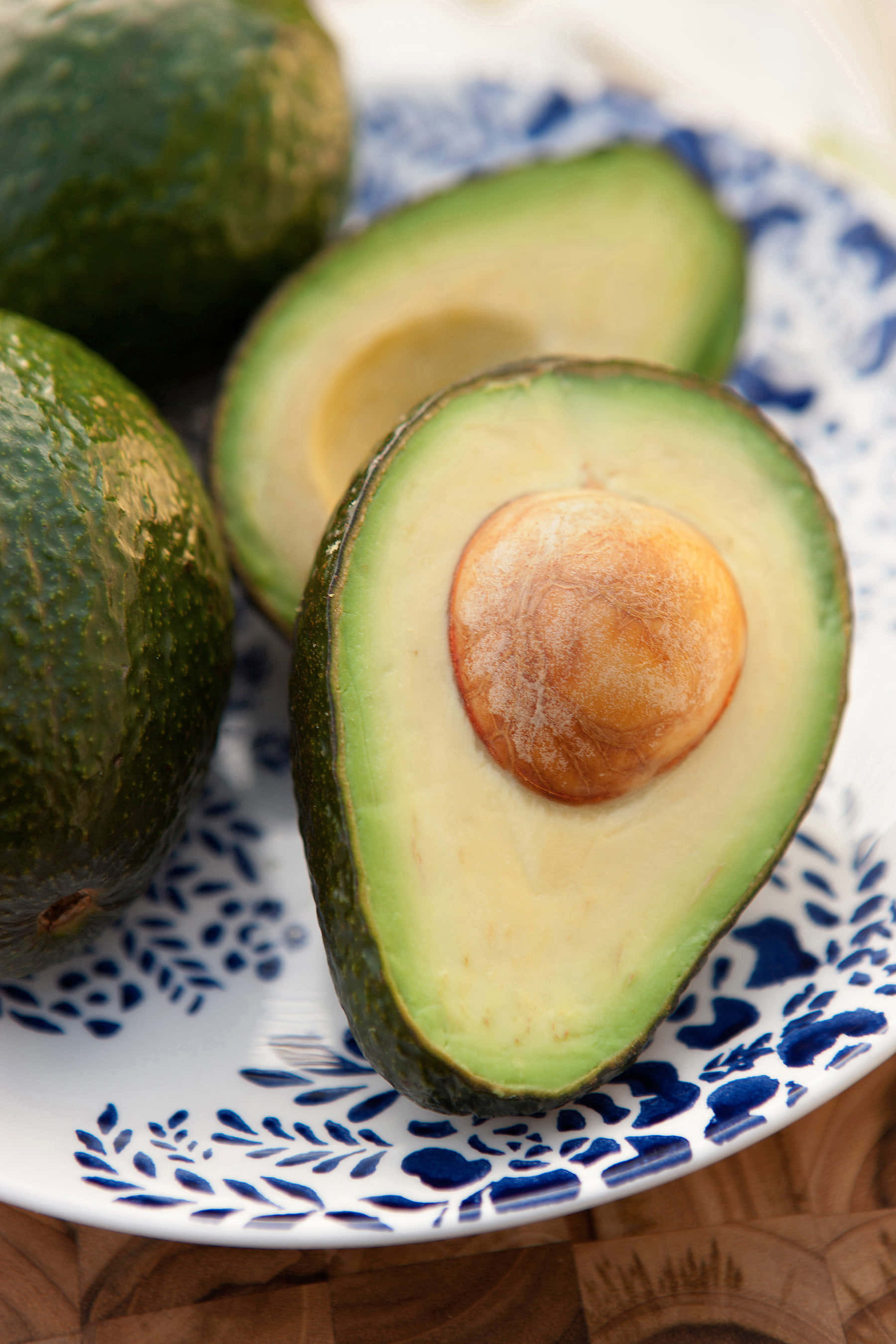 Avocado 桌布 壁紙 — 半個成熟的酪梨，帶有奶油狀的果肉和大籽，放在一個藍色圖案盤子上，旁邊有完整的酪梨。