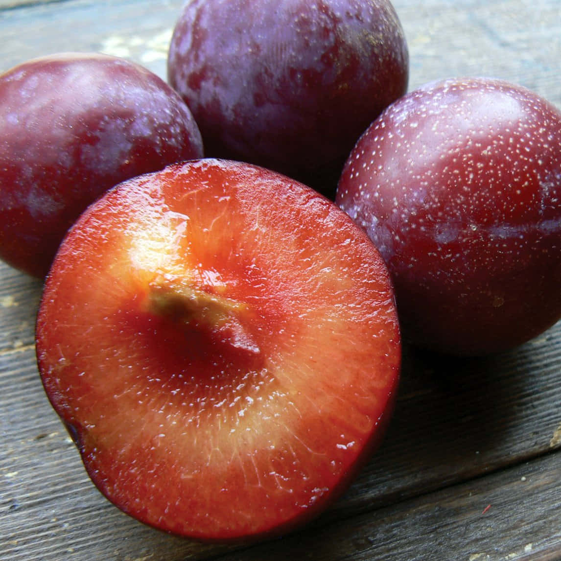 Red Plum 桌布 壁紙 — 半個李子露出多汁的紅色果肉，旁邊放著完整的李子，置於經風化的木質表面上。