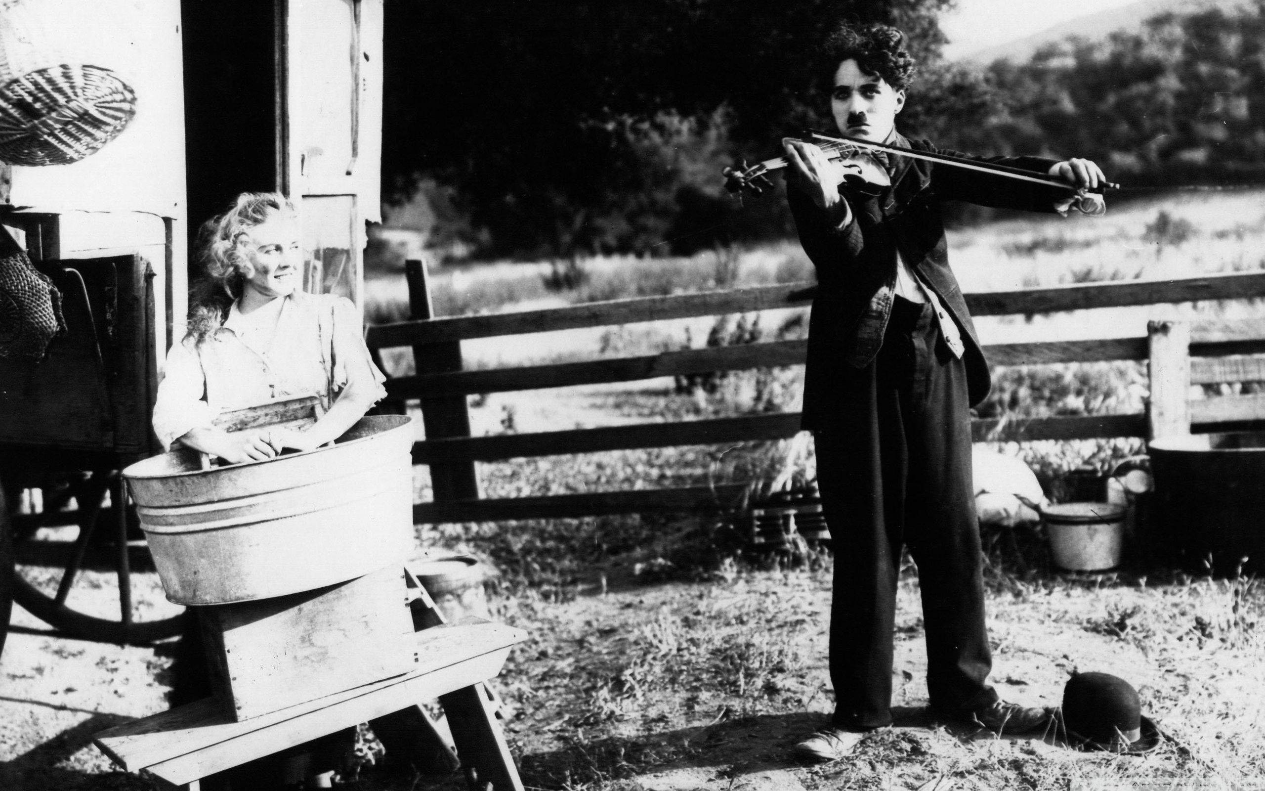 Chaplin 桌布 壁紙 — 卓別林在一個有木柵欄和樹木的簡陋小屋外，在一個洗衣盆旁邊為一位女士拉小提琴。