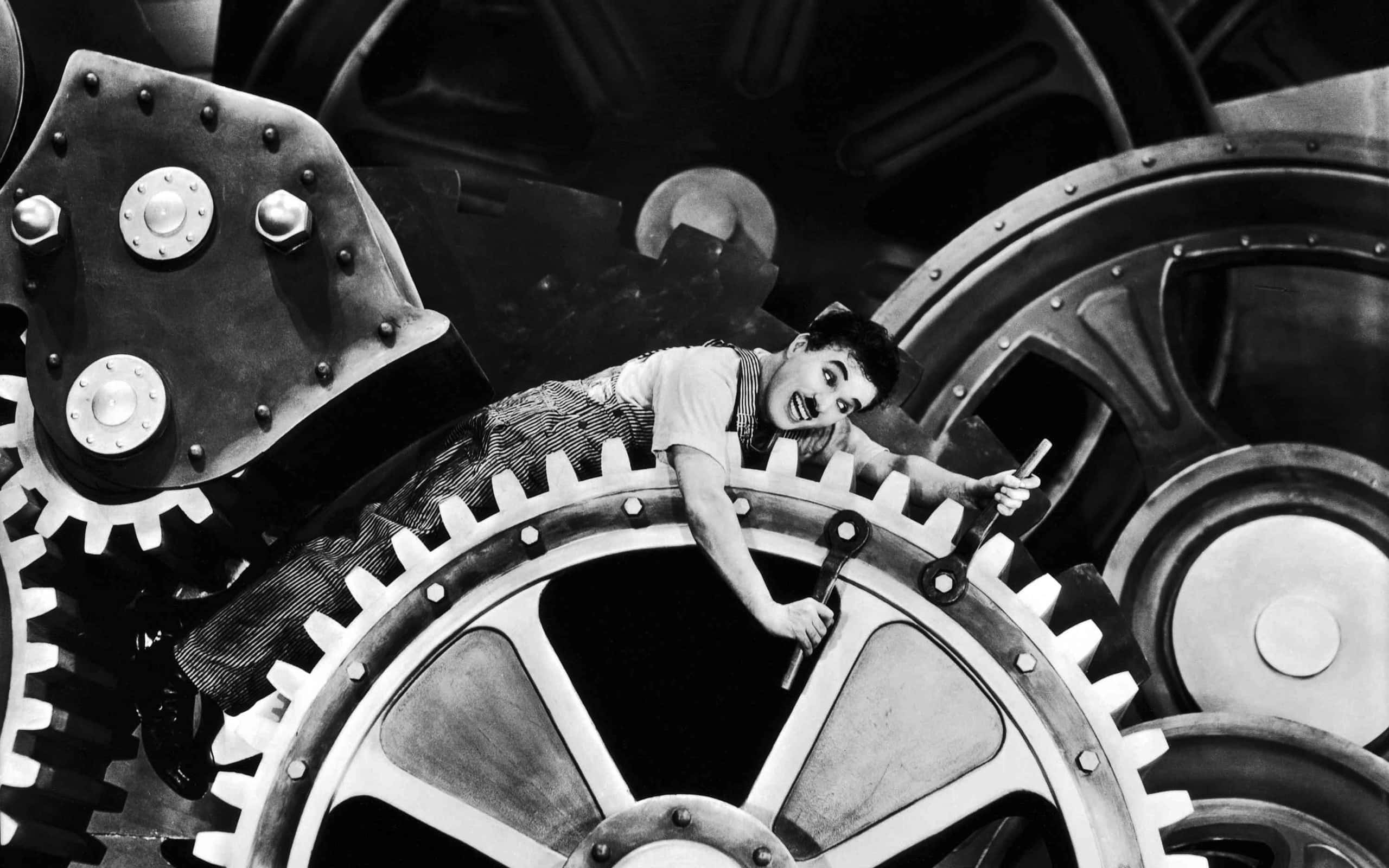 Black And White Movie 桌布 壁紙 — 卓別林在經典默片場景中，頑皮地咧嘴而笑，棲身於一個巨大的工業齒輪之上。