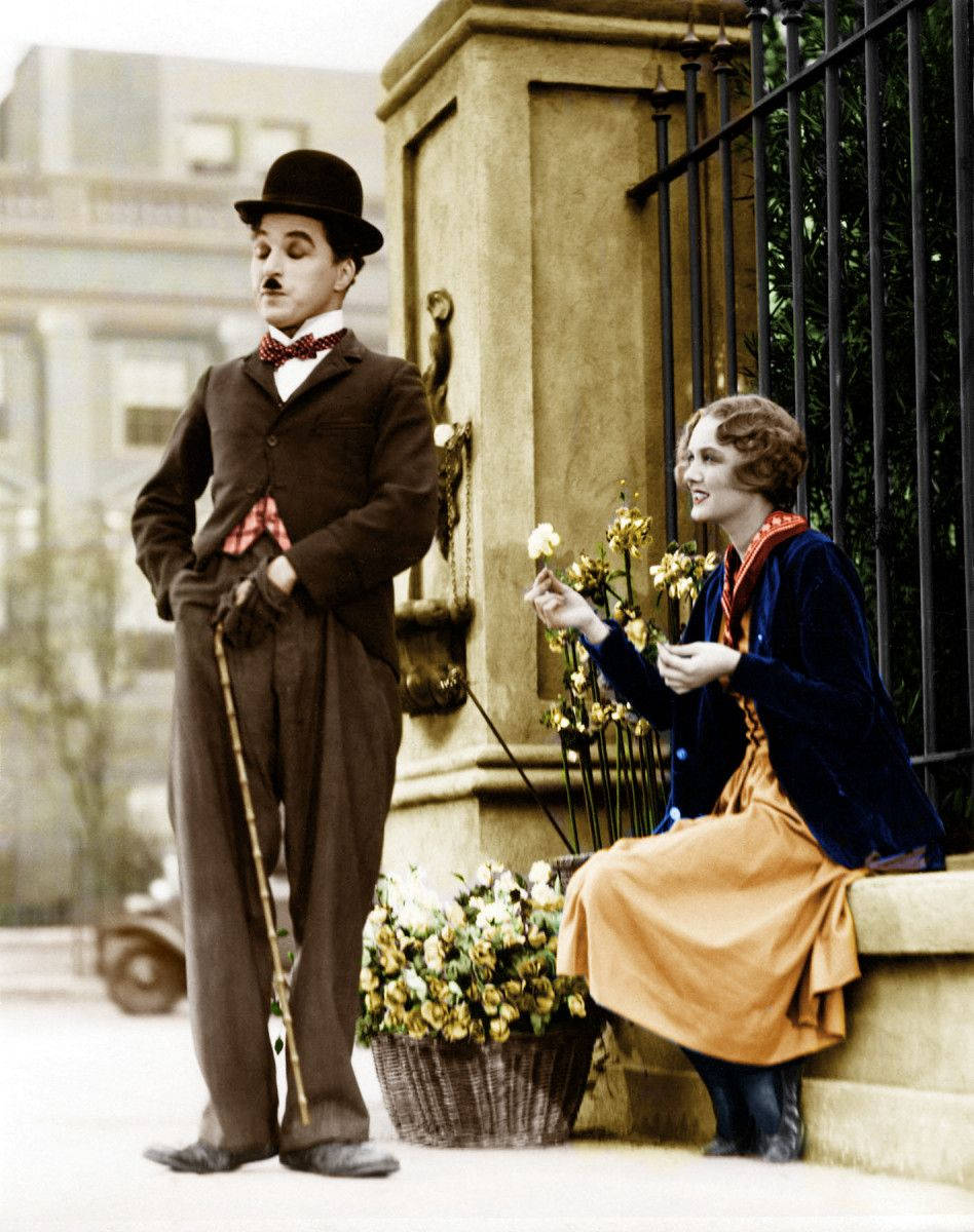Chaplin 桌布 壁紙 — 卓別林戴著圓頂硬禮帽，站在一位身穿藍色天鵝絨外套、手持鮮花的女士旁邊，靠近石門。