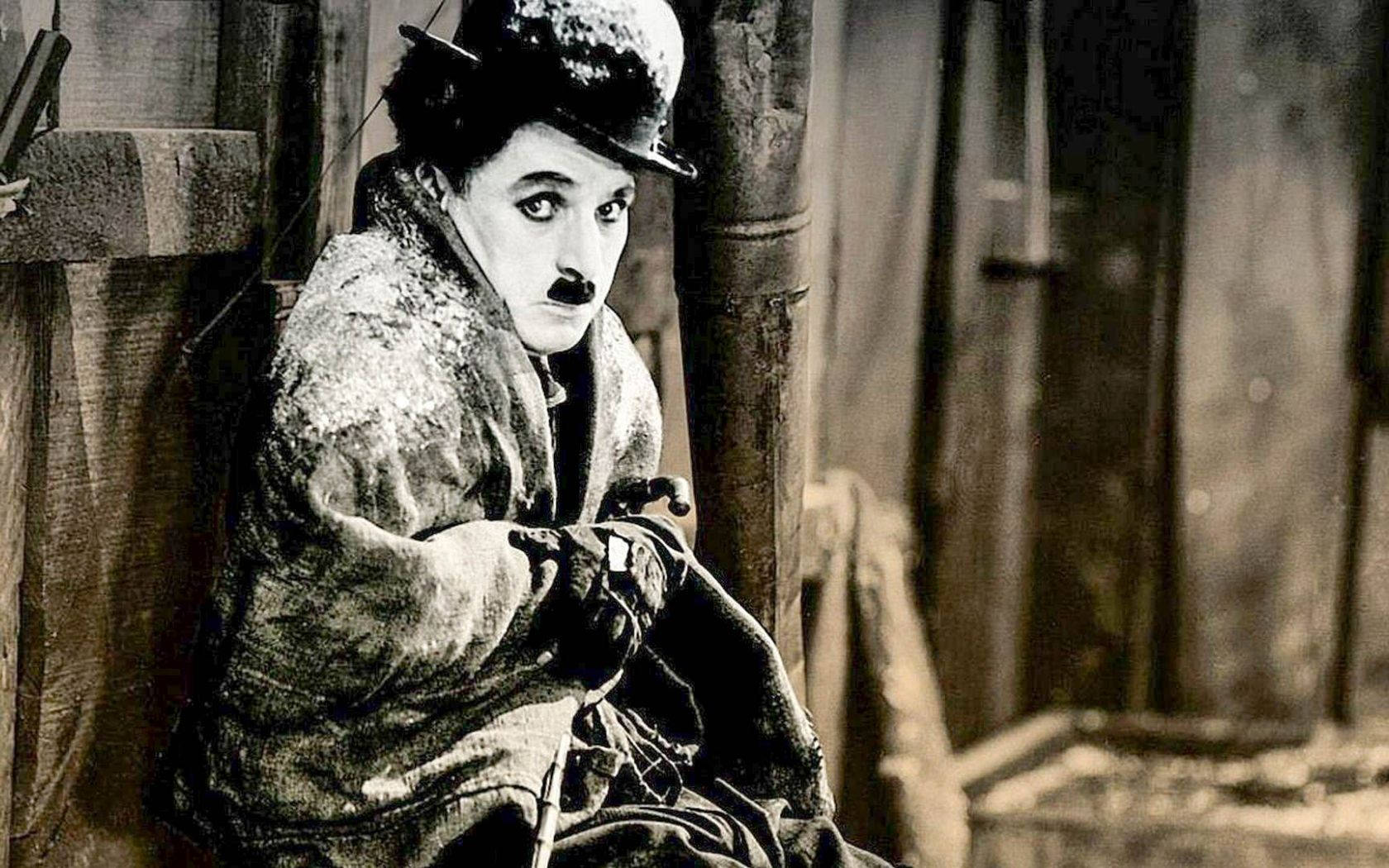 Chaplin 桌布 壁紙 — 卓別林戴著標誌性的圓頂硬禮帽和手杖，蜷縮在木板上，粗糙的黑白街景。
