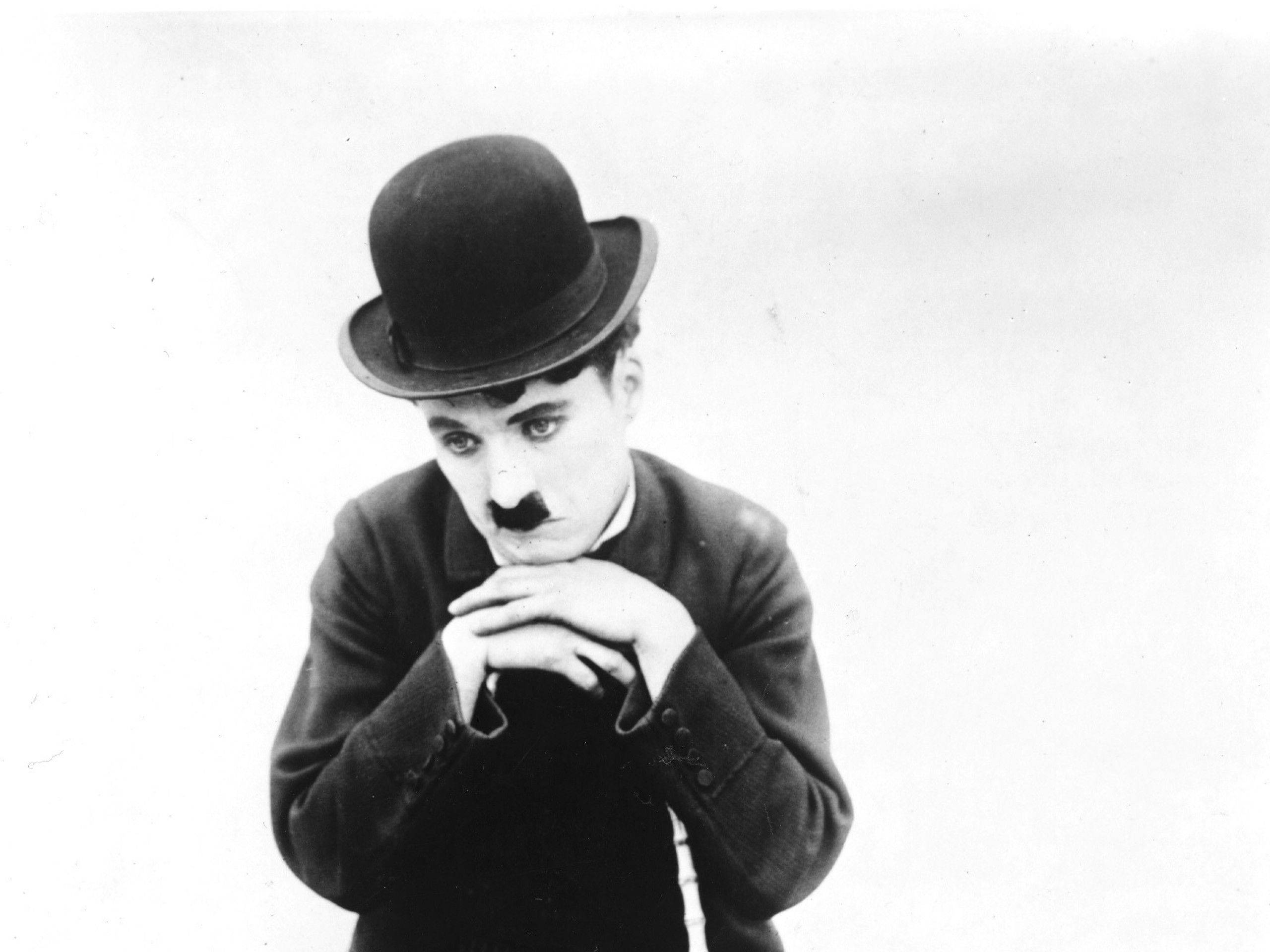 Chaplin 桌布 壁紙 — 卓別林戴著標誌性的圓頂禮帽和鬍子，雙手交握，在樸素背景下若有所思地凝視。