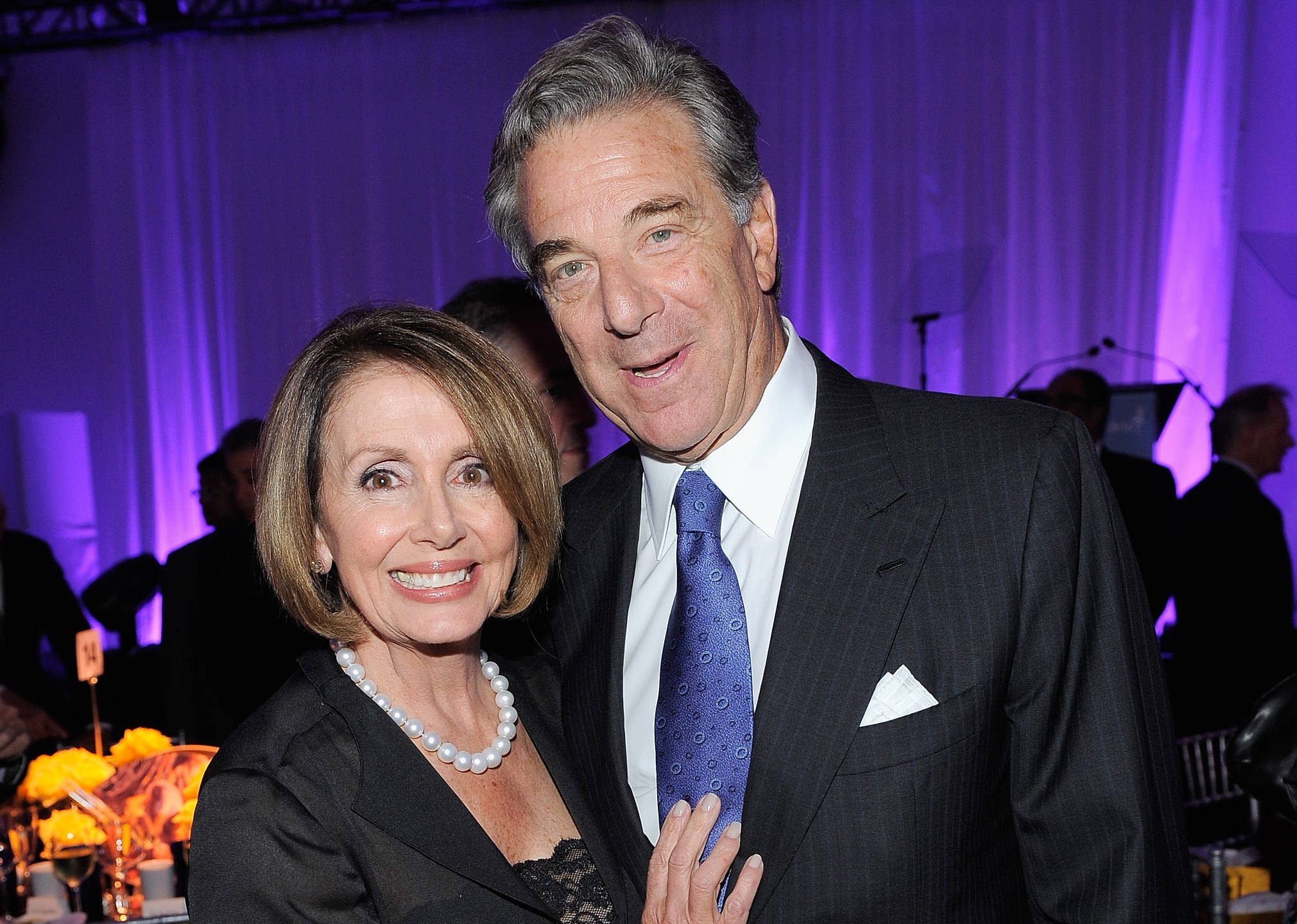 Nancy Pelosi 桌布 壁紙 — 南希·佩洛西和一位穿西裝的男子在一個有紫色燈光和花卉桌子的正式活動中溫暖地微笑。