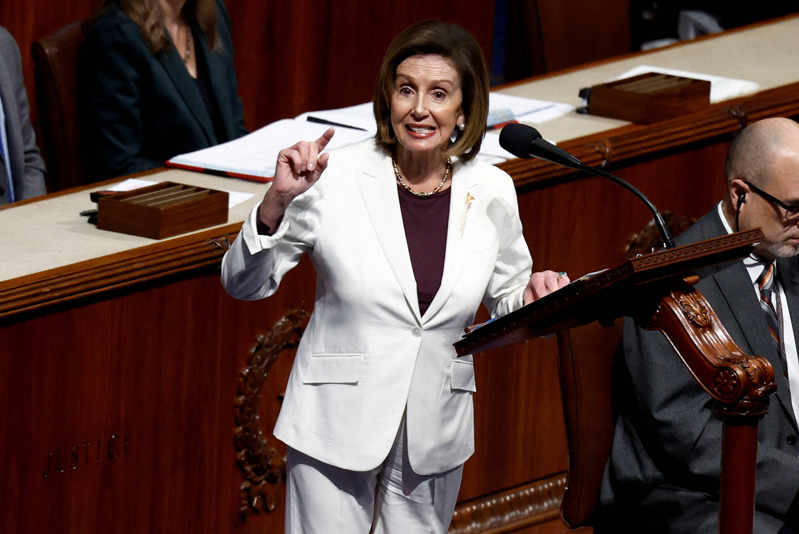 Nancy Pelosi 桌布 壁紙 — 南希·裴洛西在一個裝飾華麗木工、正式的立法會議廳內，於木製講台上充滿激情地發言，附近有專注的工作人員。