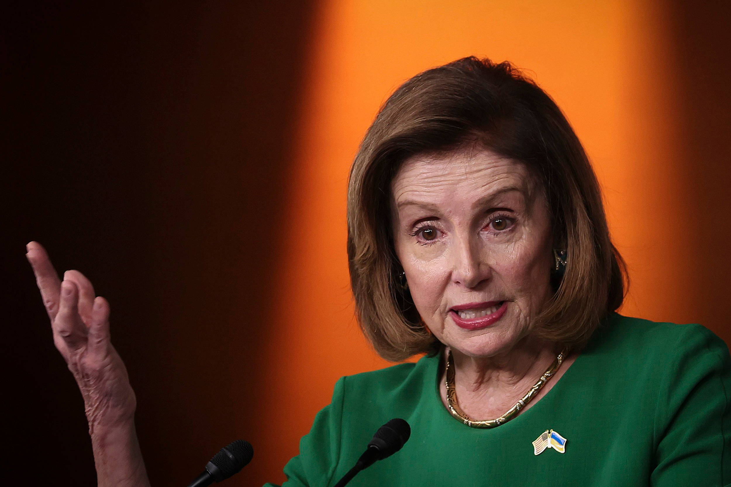 Nancy Pelosi 桌布 壁紙 — 南希·裴洛西在溫暖的橙色背景下，於講台上激情演講，身穿綠色西裝並佩戴烏克蘭國旗別針。