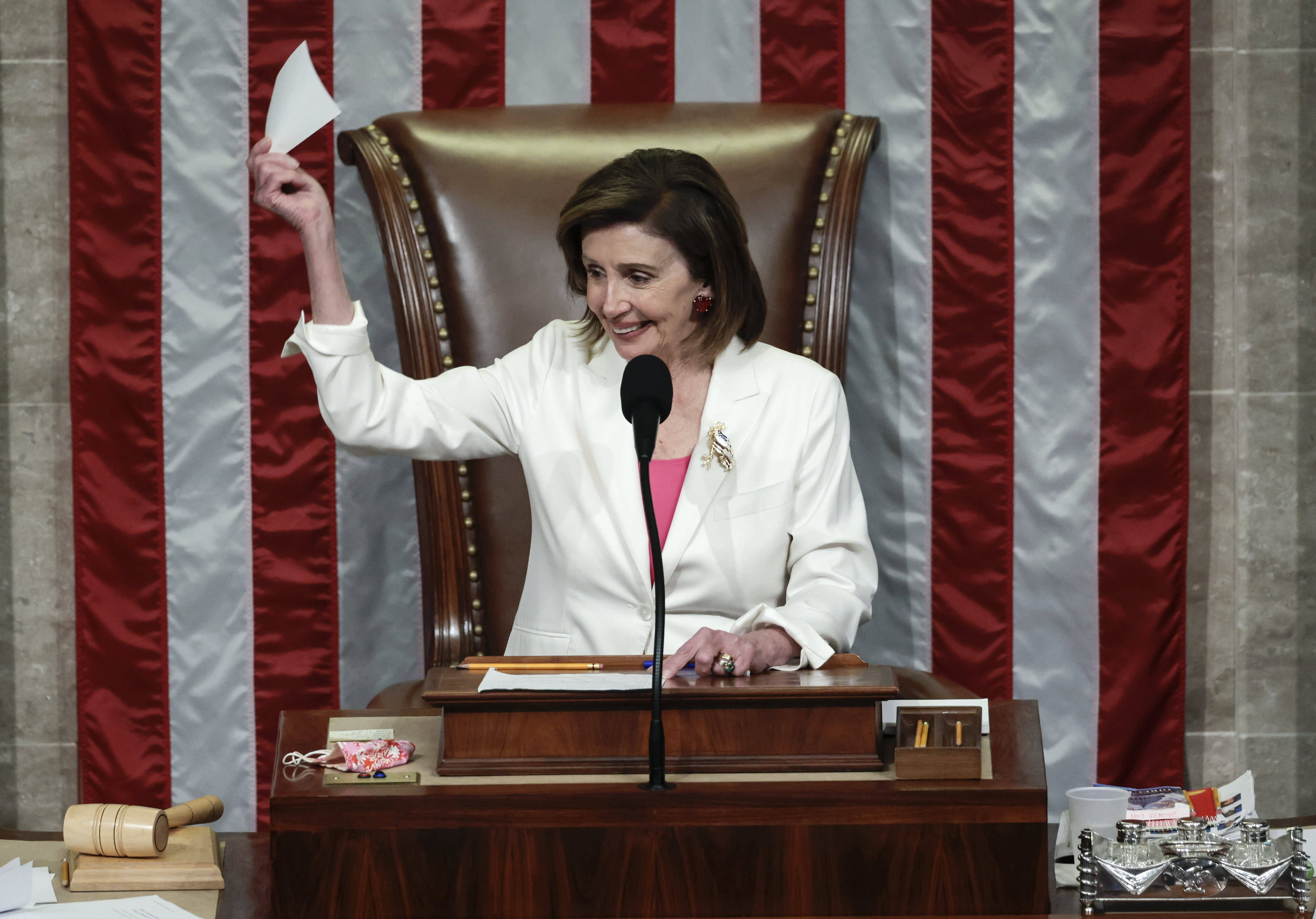 Nancy Pelosi 桌布 壁紙 — 南希·裴洛西在美國國會大廈的辦公桌前微笑著舉起一份文件，背後有美國國旗條紋。