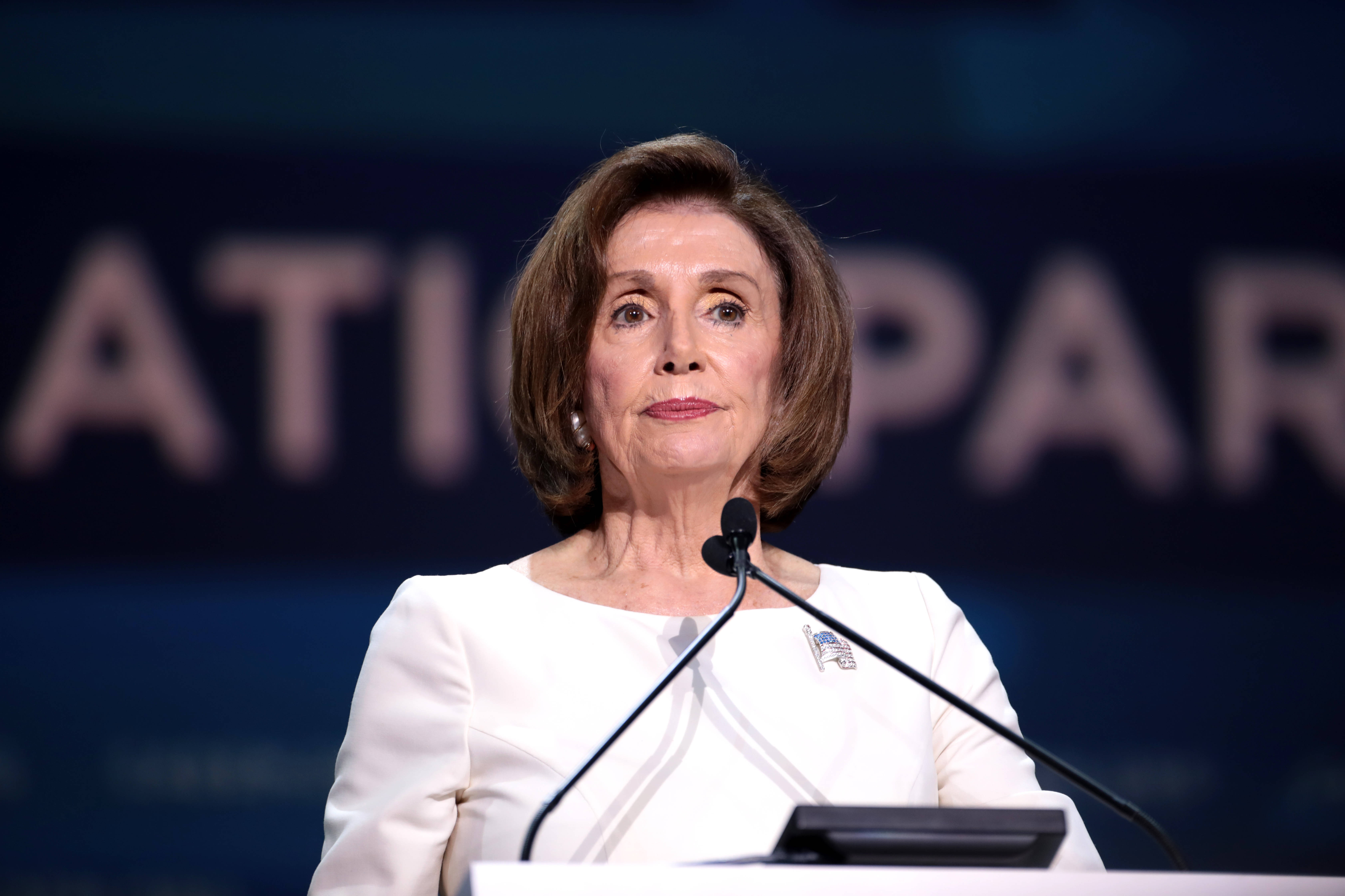 Nancy Pelosi 桌布 壁紙 — 南希·裴洛西站在講台上發表嚴肅演講，背景為深藍色並帶有模糊文字。