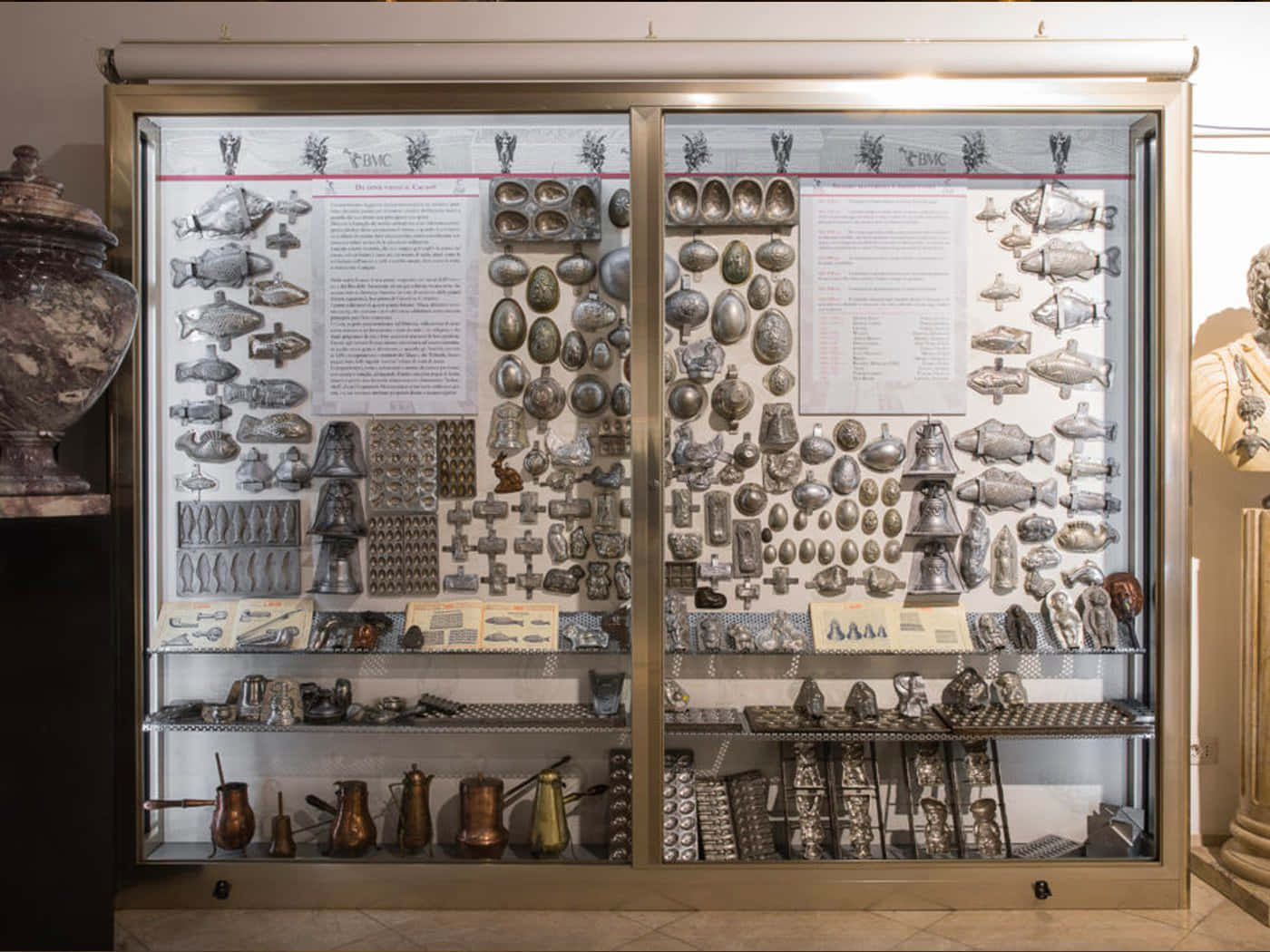Italian 桌布 壁紙 — 博物館展示櫃內放滿精緻的銀魚模具、蛋模具和銅鍋，在明亮燈光下展示。