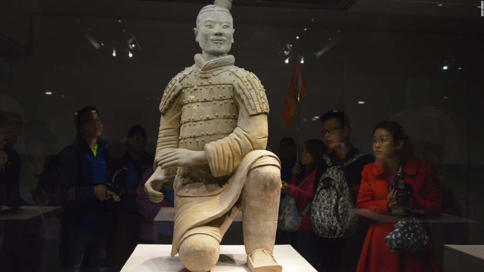 Museum 桌布 壁紙 — 博物館展覽中一個跪著的兵馬俑雕像，背景有參觀者在觀察。