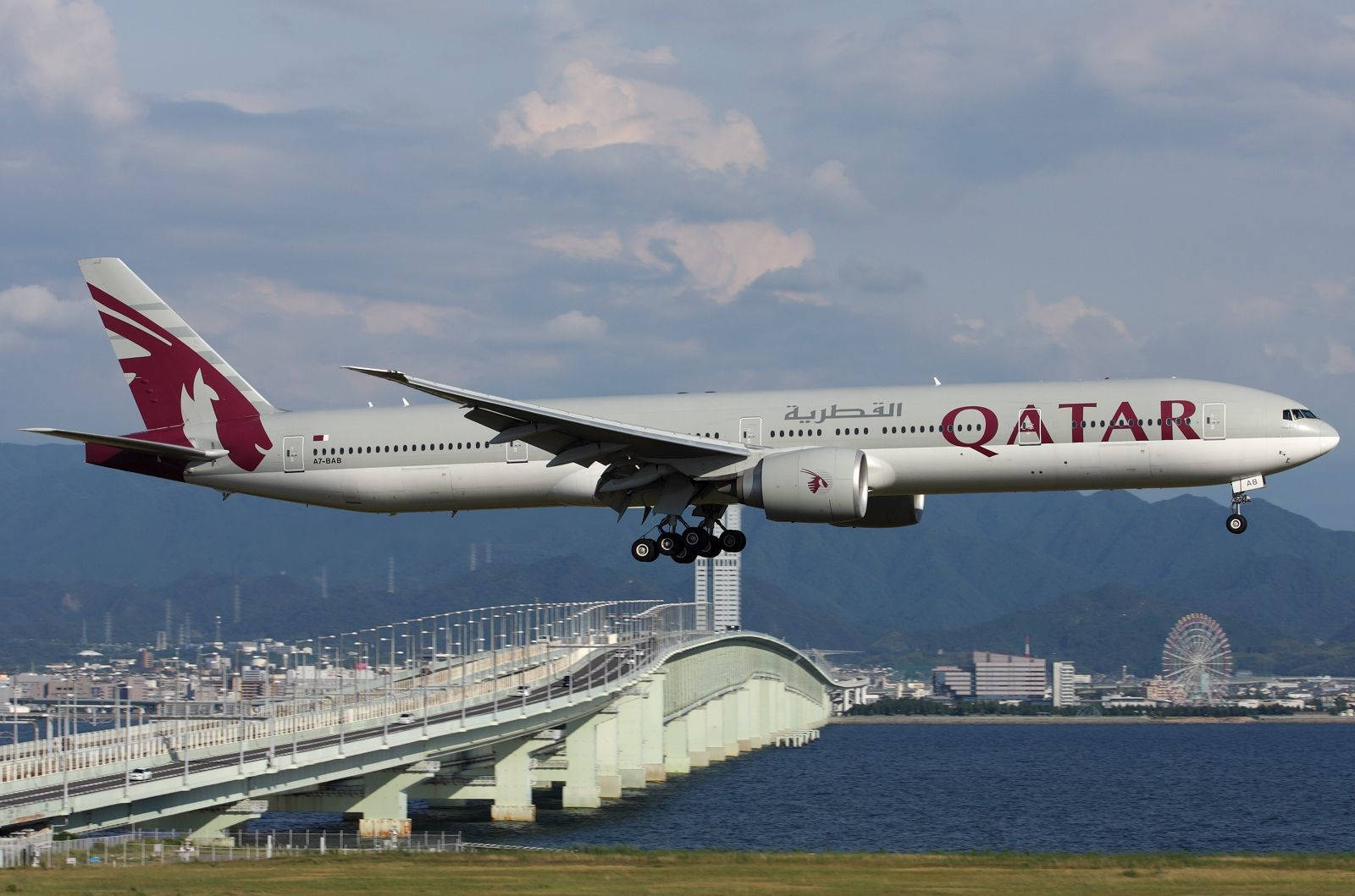 飛機 桌布 壁紙 — 卡塔爾航空波音777在陰天背景下，於一座橋樑上空起飛，背景有城市天際線和摩天輪。
