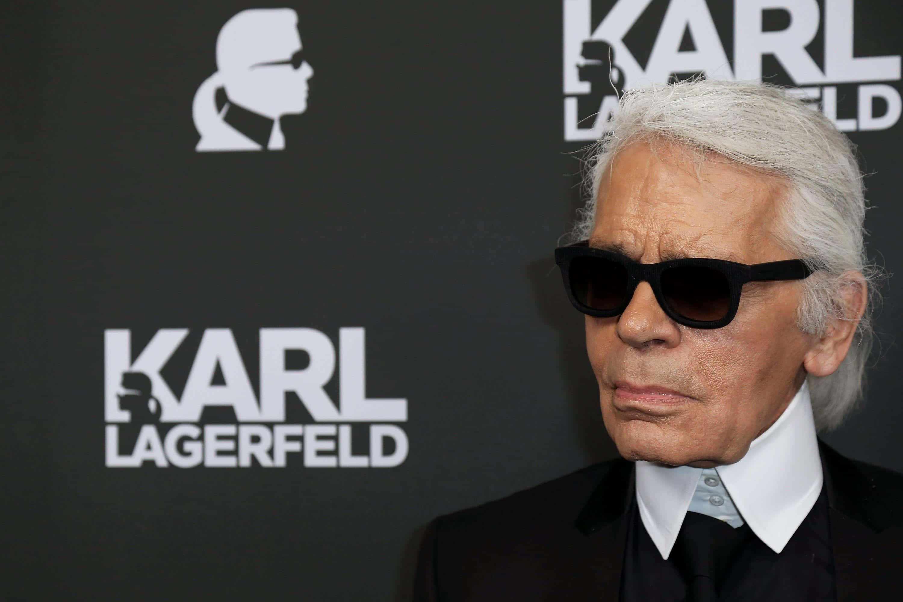 Karl Lagerfeld 桌布 壁紙 — 卡爾·拉格斐戴著黑色太陽眼鏡和西裝，站在黑色背景前，背景上有他的名字和剪影標誌。