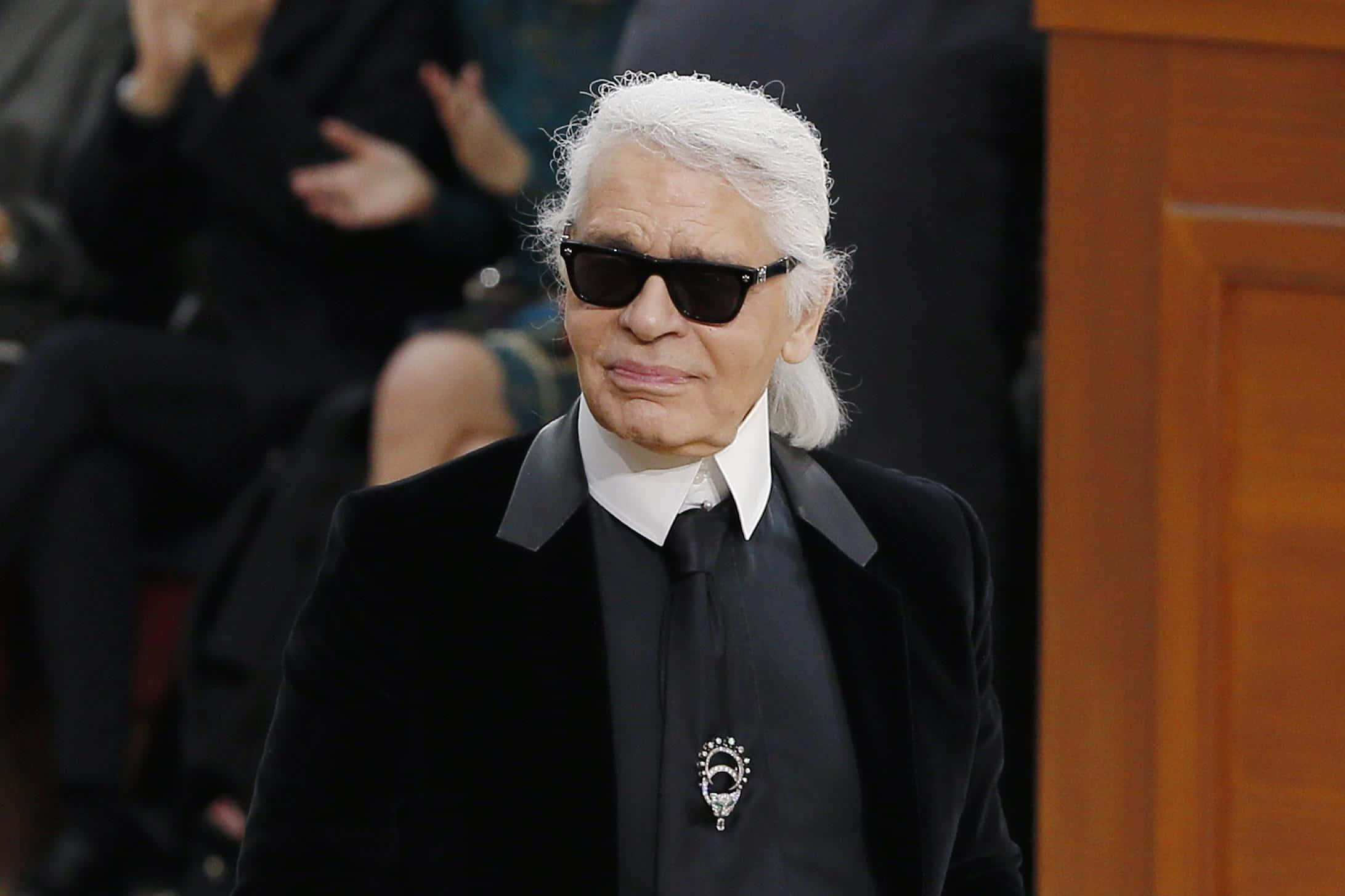 Karl Lagerfeld 桌布 壁紙 — 卡爾·拉格斐穿著黑色西裝和太陽眼鏡，表情嚴肅地走在時尚活動中。