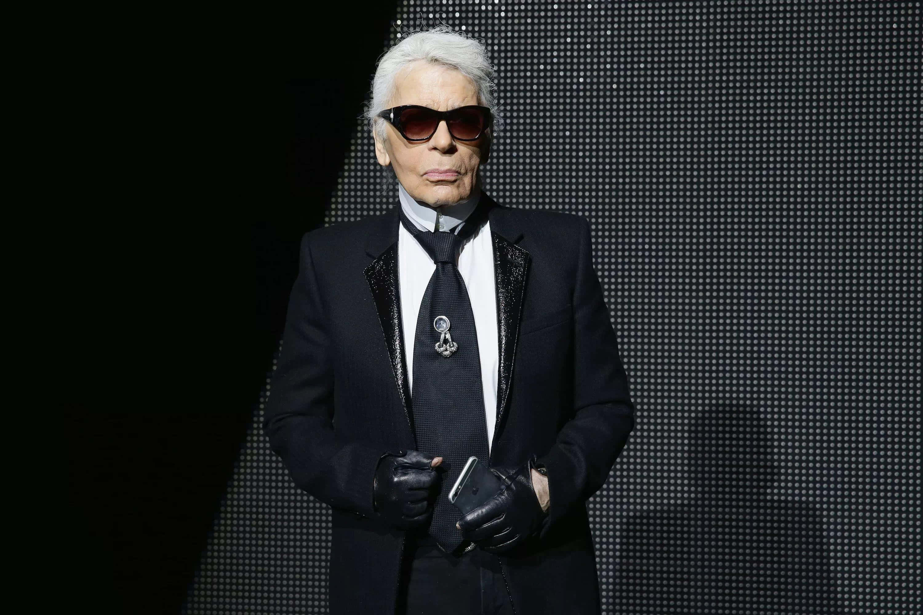 Karl Lagerfeld 桌布 壁紙 — 卡爾·拉格斐自信地穿著黑色西裝和太陽眼鏡，站在深色像素背景前。