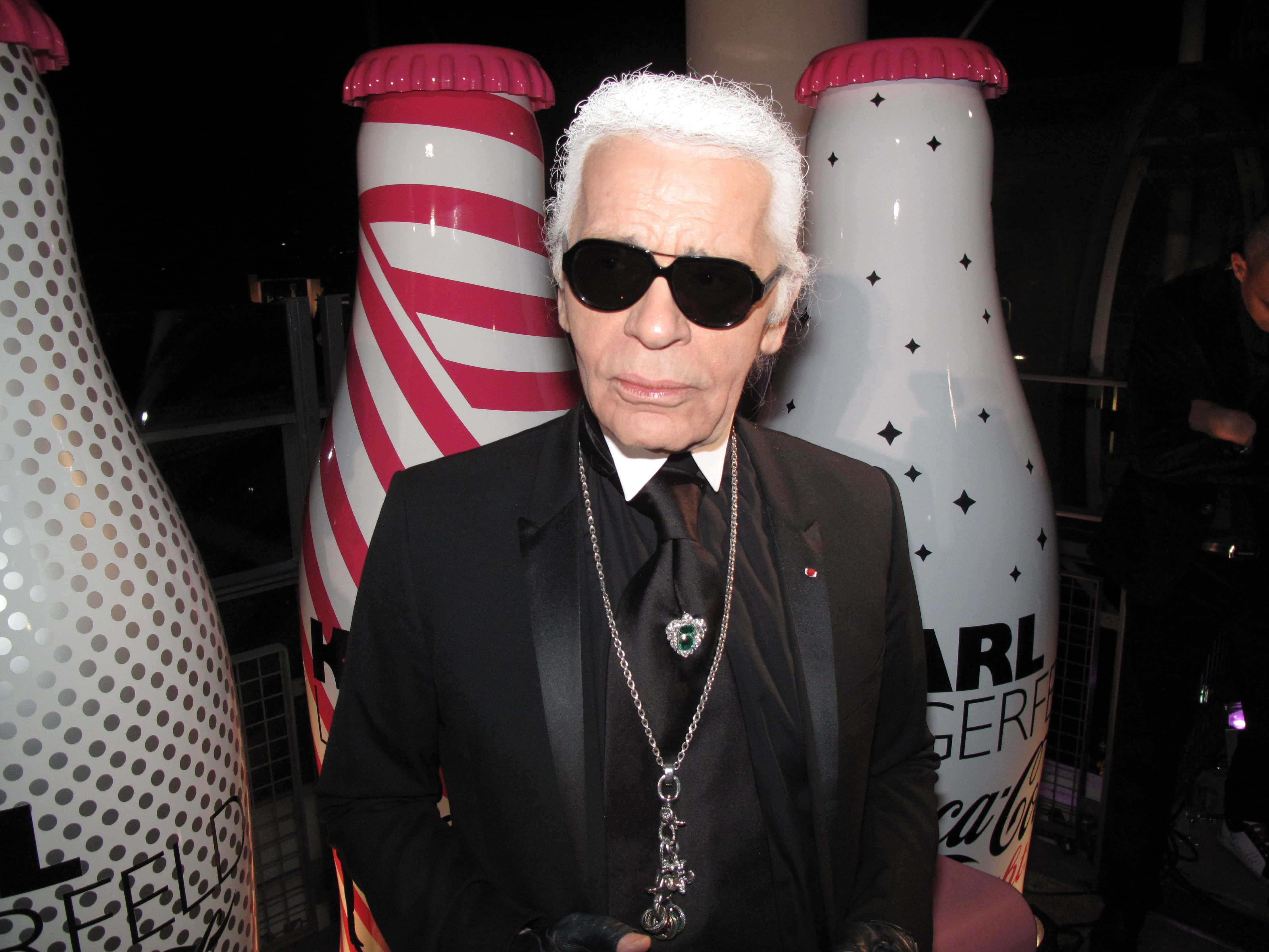 Karl Lagerfeld 桌布 壁紙 — 卡爾·拉格斐身穿黑色西裝和太陽眼鏡，在夜間活動中站在巨大的風格化可口可樂瓶前。