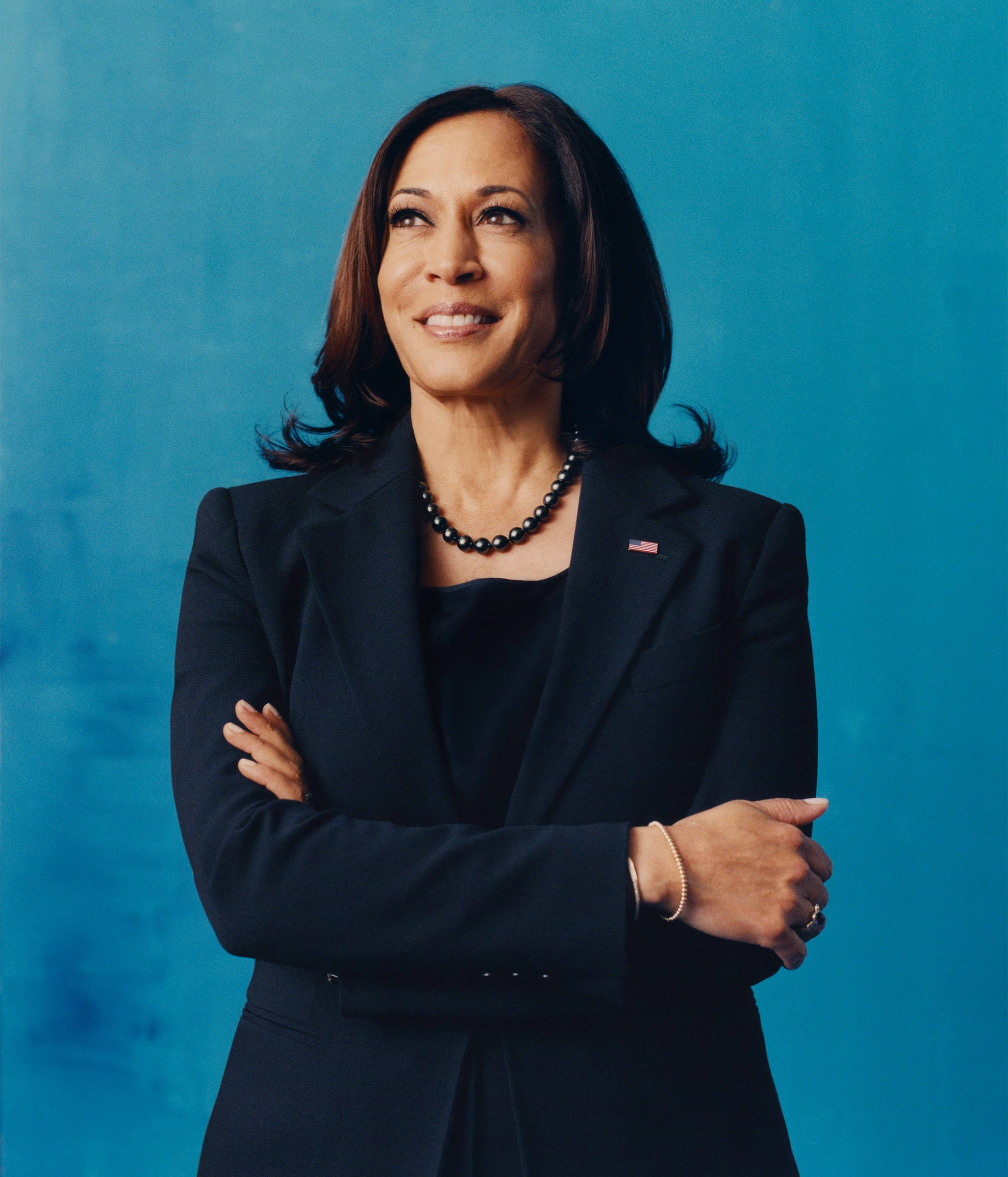 Kamala Harris 桌布 壁紙 — 卡瑪拉·哈里斯自信地交叉雙臂，站在純藍色背景前，戴著珍珠項鍊和美國國旗別針，溫暖地微笑。