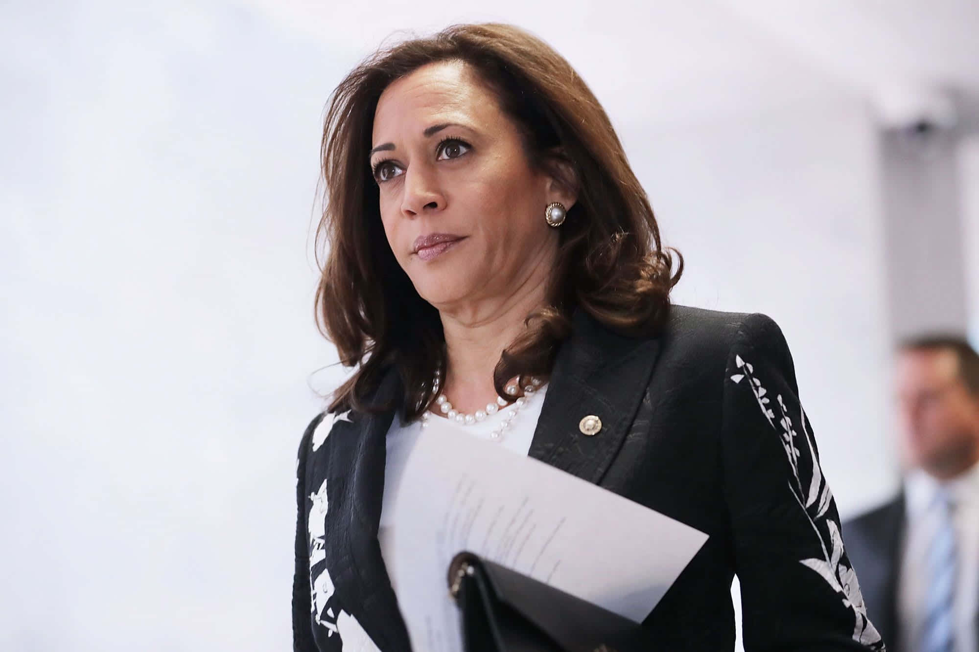 Kamala Harris 桌布 壁紙 — 卡瑪拉·哈里斯身穿黑色花卉西裝外套，在明亮的室內環境中手持文件，表情嚴肅。