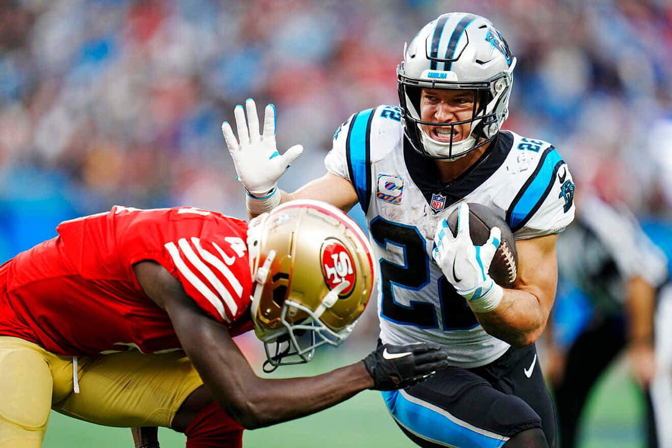 Christian Mccaffrey 桌布 壁紙 — 卡羅萊納黑豹隊的跑衛身穿白色和藍色裝備，緊抱著橄欖球，同時被身穿紅色和金色的舊金山49人隊防守球員擒抱。