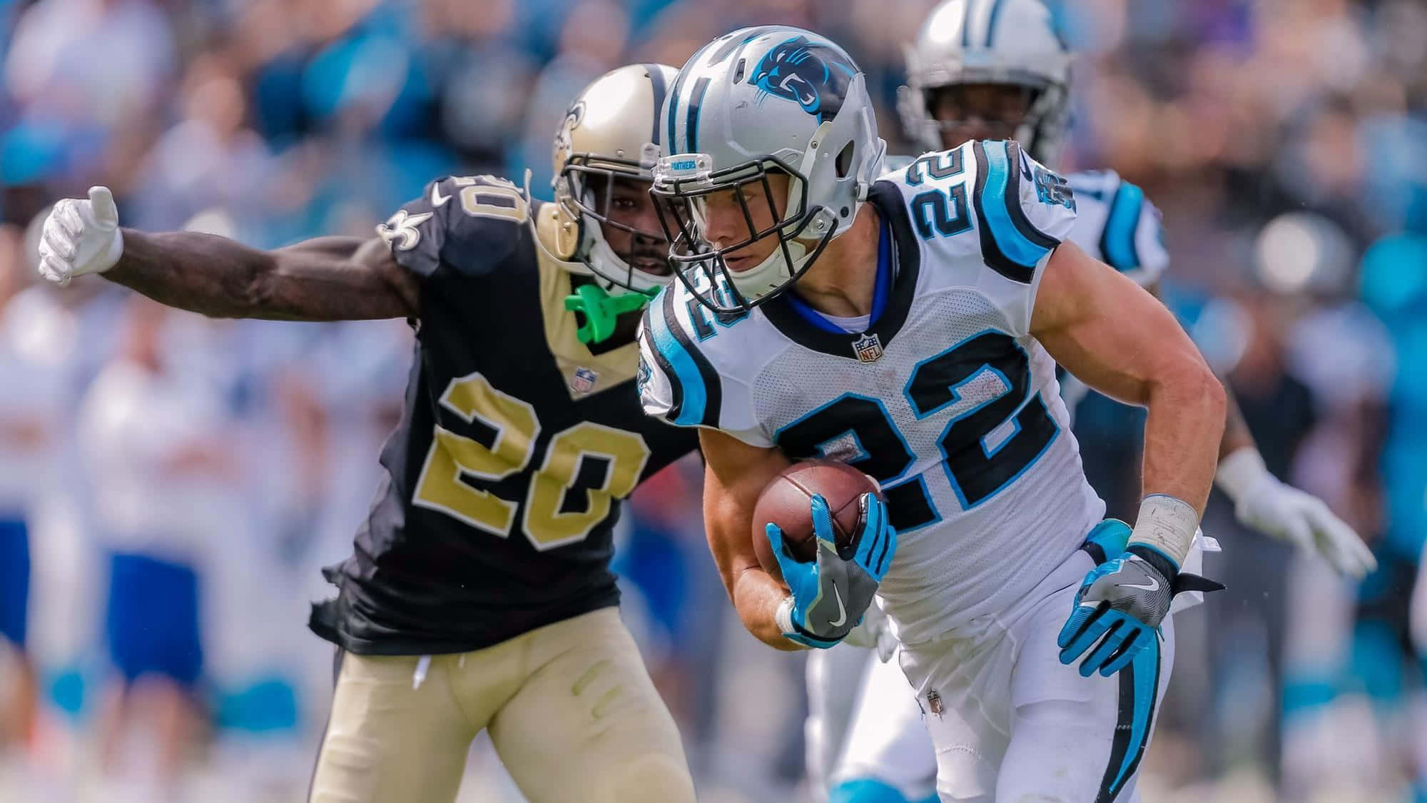Christian Mccaffrey 桌布 壁紙 — 卡羅萊納黑豹隊的跑衛身穿白色和藍色裝備，帶著橄欖球衝刺，而紐奧良聖徒隊的防守球員身穿黑色和金色在日間NFL比賽中逼近。