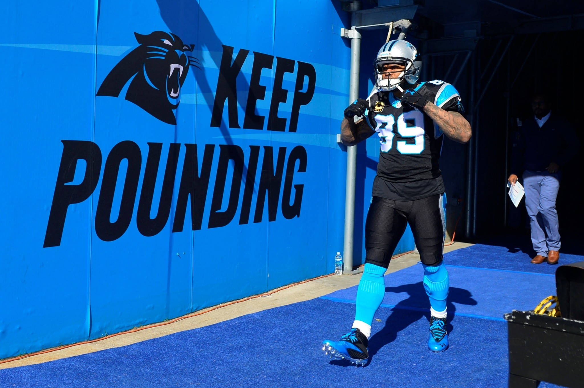 Panthers 桌布 壁紙 — 卡羅萊納黑豹隊美式足球員身穿黑藍裝備，自信地走過藍色牆壁，牆上有「KEEP POUNDING」和咆哮的黑豹標誌。
