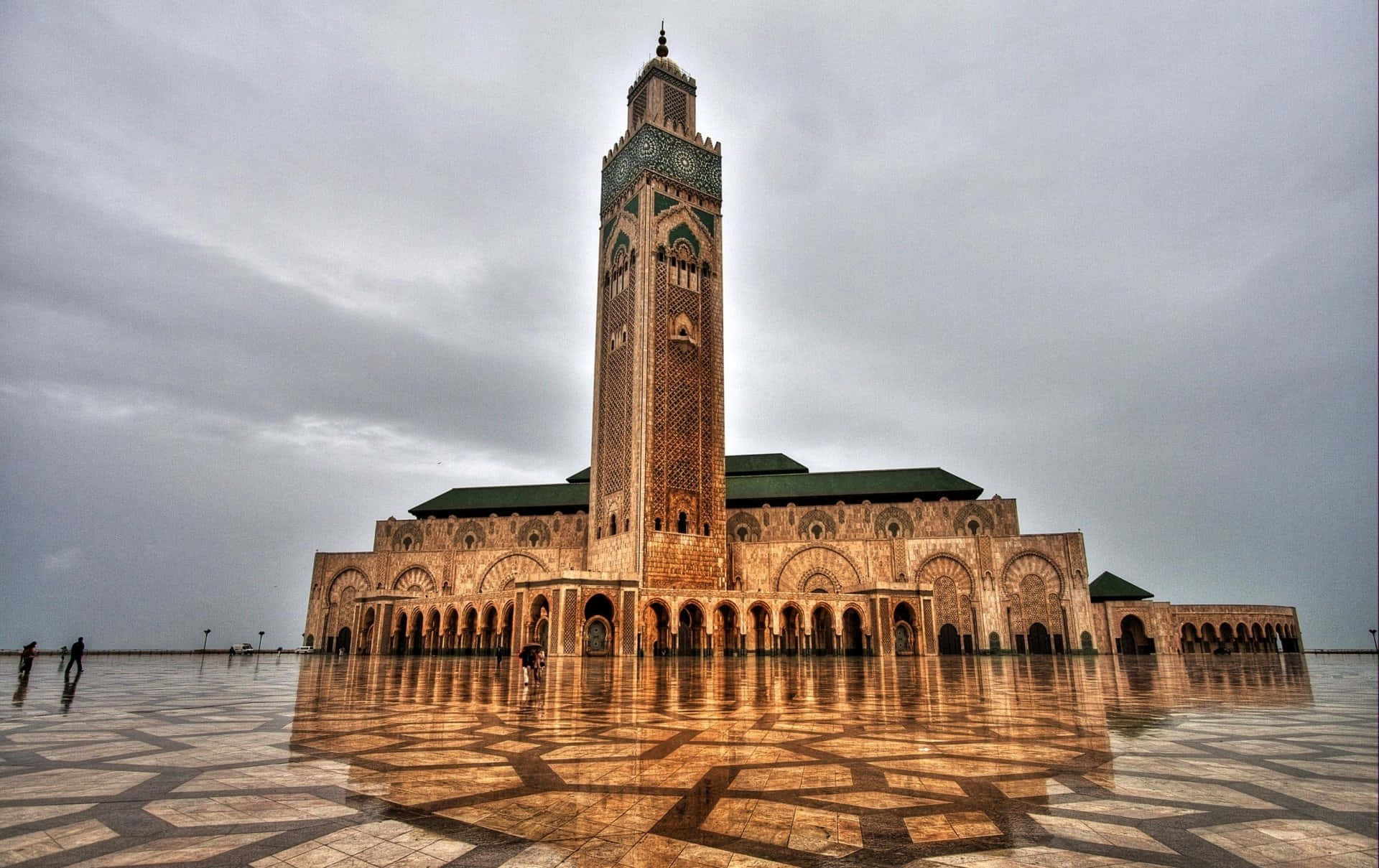 Mosque 桌布 壁紙 — 卡薩布蘭卡的哈桑二世大清真寺在陰天天空下巍然聳立，濕潤的瓷磚廣場映照出其精緻的建築。