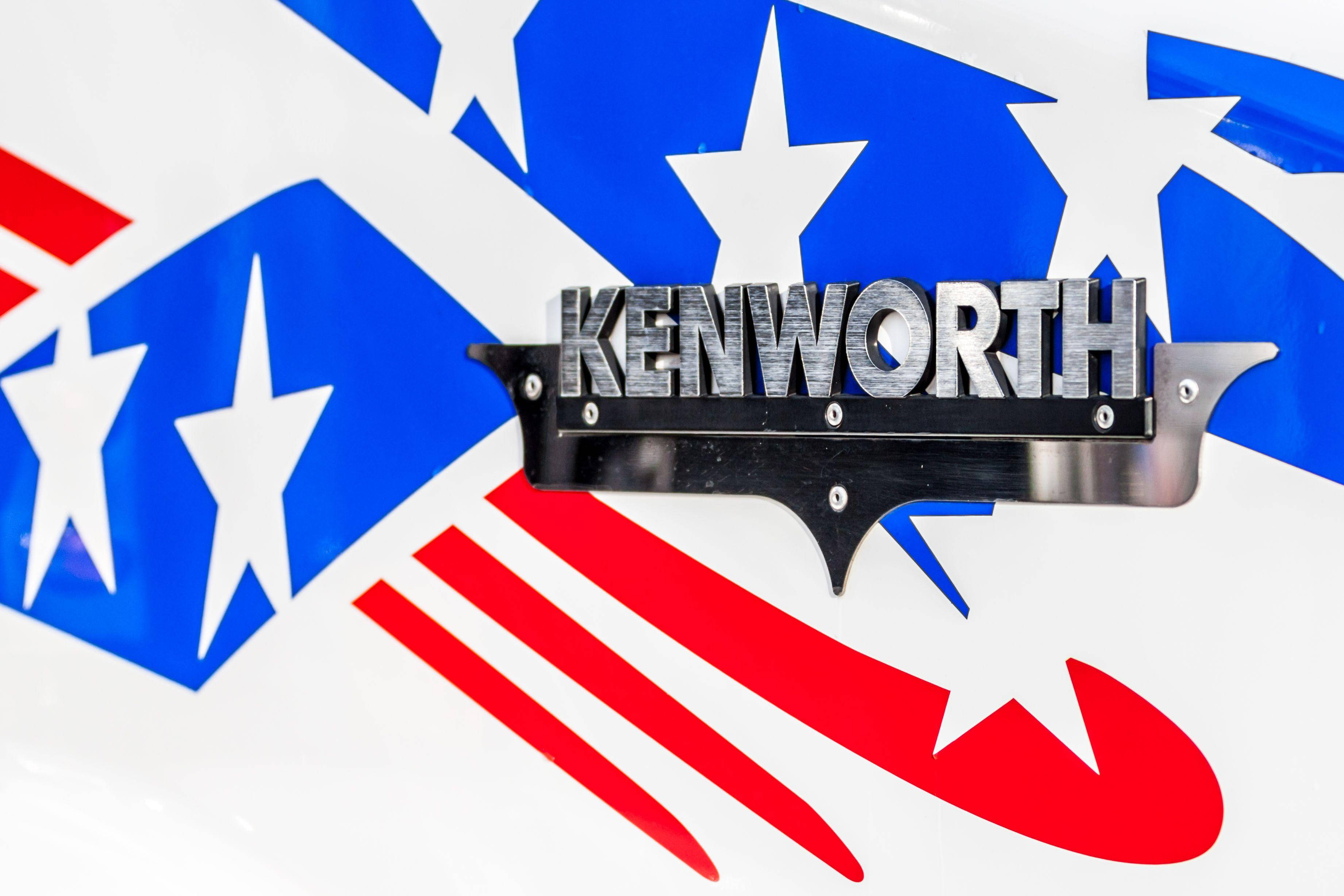 肯沃斯 桌布 壁紙 — 卡車上安裝的鍍鉻 Kenworth 標誌特寫，帶有醒目的紅、白、藍星條紋圖案。