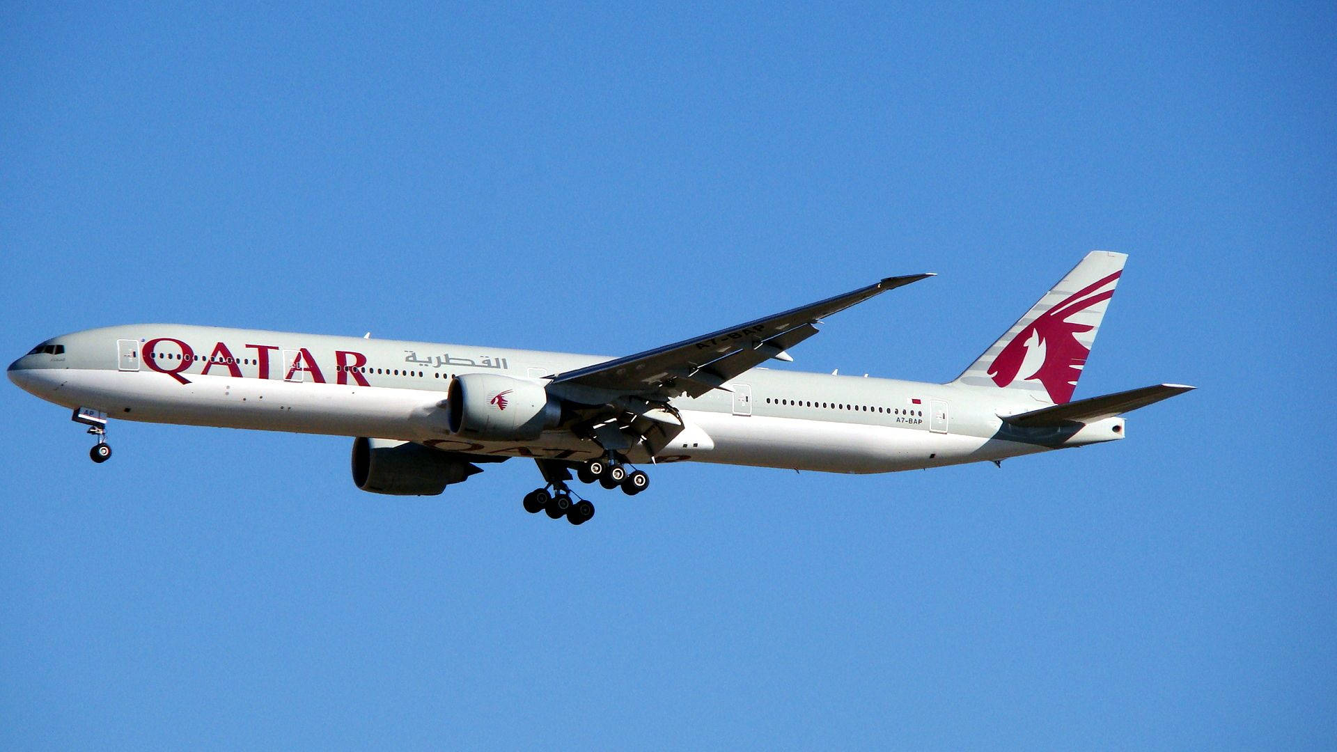 飛機 桌布 壁紙 — 卡達航空波音777以白色和栗色塗裝，在白天飛行中優雅地傾斜，映襯著清澈的藍天。