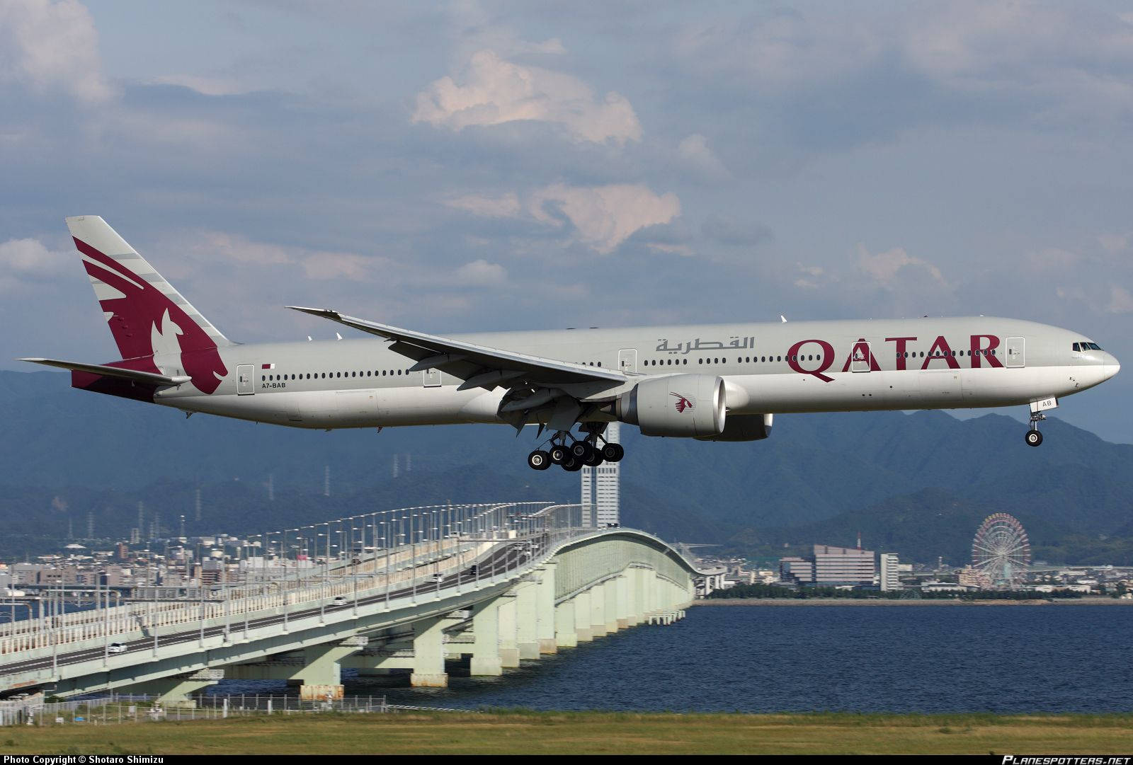 Qatar 桌布 壁紙 — 卡達航空波音777在沿海橋樑上空低飛起飛，背景有山脈和城市景觀。
