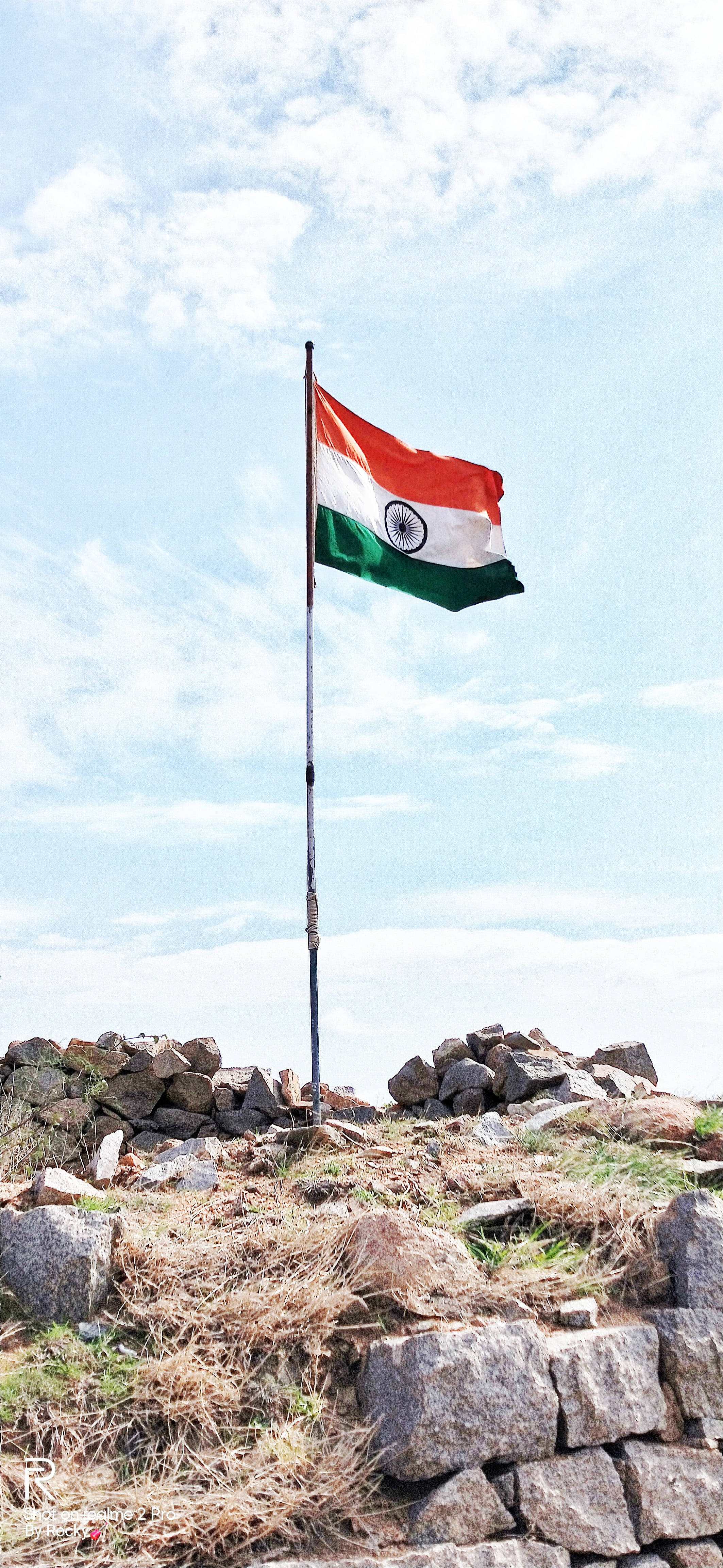 India 桌布 壁紙 — 印度國旗在岩石山頂上，在晴朗藍天和零星白雲下自豪地飄揚。