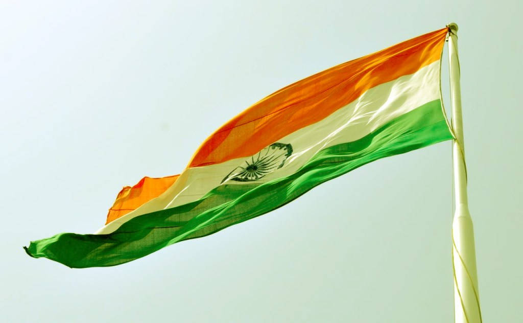 Indian 桌布 壁紙 — 印度國旗在淡色天空下動態飄揚，帶有藏紅花、白色和綠色條紋，中央為阿育王法輪。