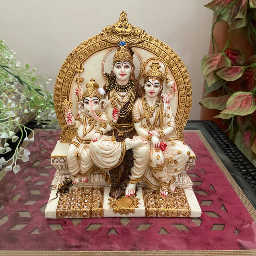 Shiv Parivar 桌布 壁紙 — 印度教神祇濕婆帕爾瓦蒂和象頭神甘尼許的詳細雕像，一起坐在裝飾華麗的金色寶座上，帶有複雜雕刻和紅色花卉點綴。