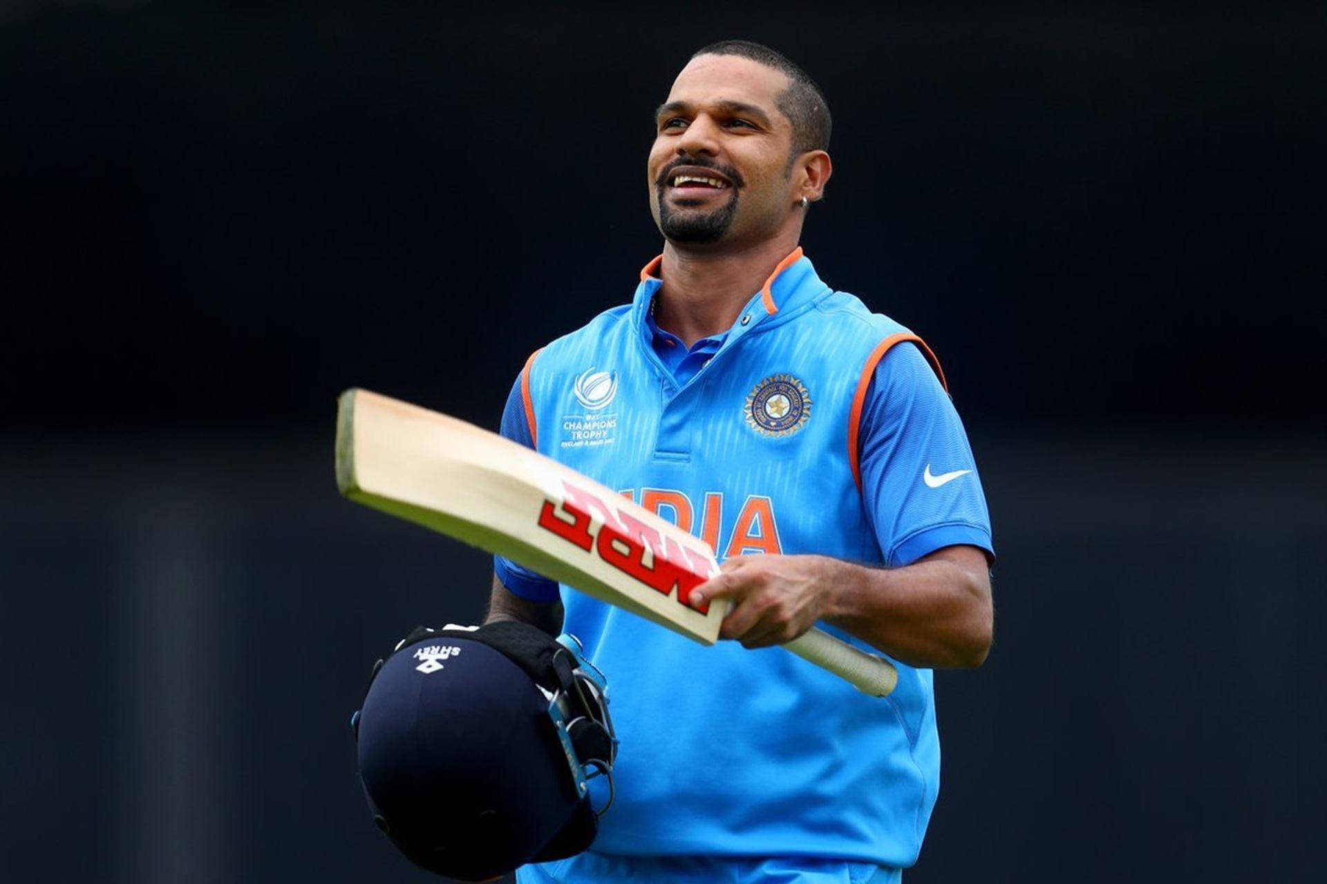 shikhar dhawan 桌布 壁紙 — 印度板球手在深色背景下自信地微笑，手持球棒和頭盔，身穿藍色隊服。