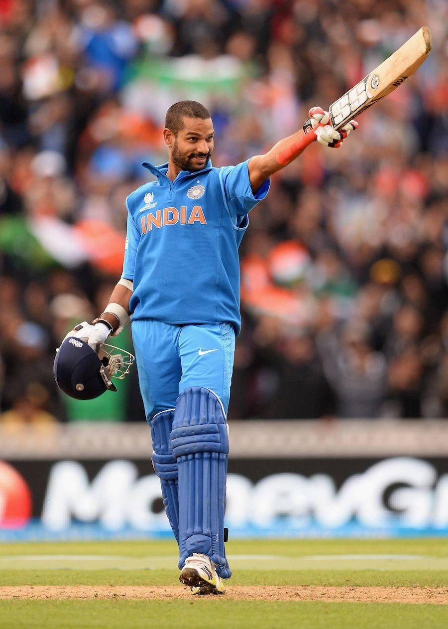 shikhar dhawan 桌布 壁紙 — 印度板球運動員在擠滿觀眾的球場上，於明亮的日光下高舉球棒慶祝。