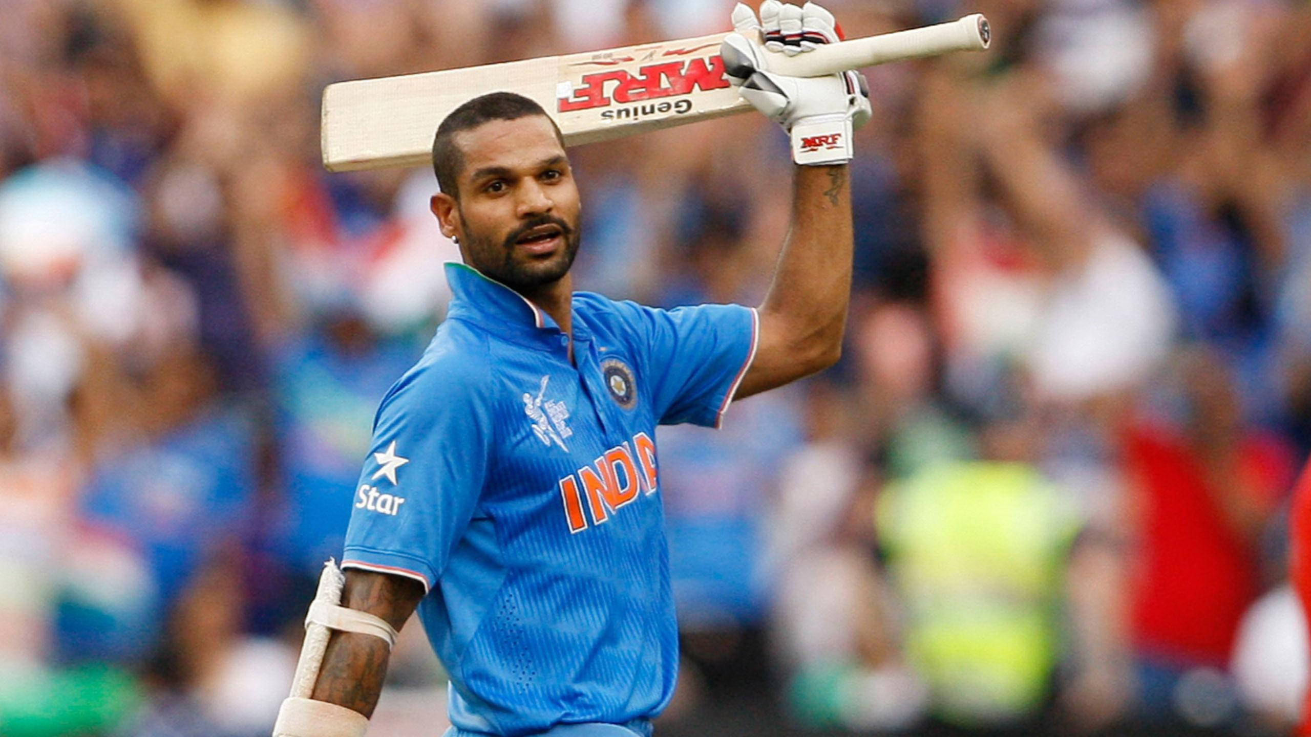 shikhar dhawan 桌布 壁紙 — 印度板球運動員在日間比賽中高舉球棒，以勝利姿態站在歡呼的人群中。